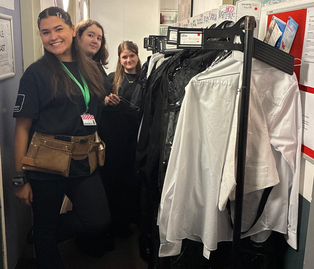 A group of three women stands in a narrow space, with two of them positioned beside a rack of clothing. One woman, wearing a black t-shirt and a green lanyard, smiles at the camera, while the others are dressed in black and look towards the side. The background shows a bulletin board and corridor walls.