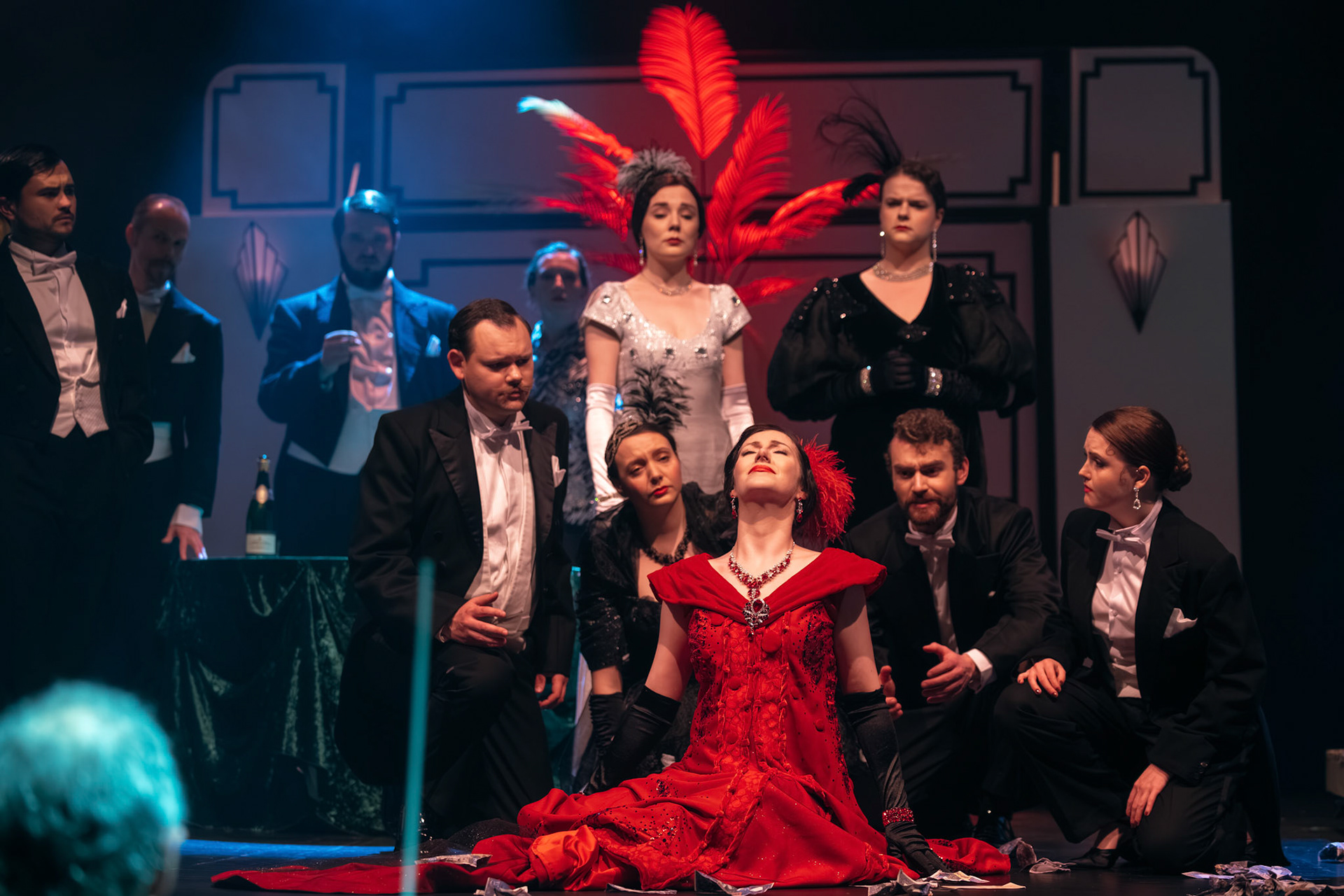 A theatrical performance featuring a woman in a red dress with a feather headdress, surrounded by a cast in formal attire. The background includes stage elements like large windows and decorative features, with dim lighting enhancing the dramatic scene.