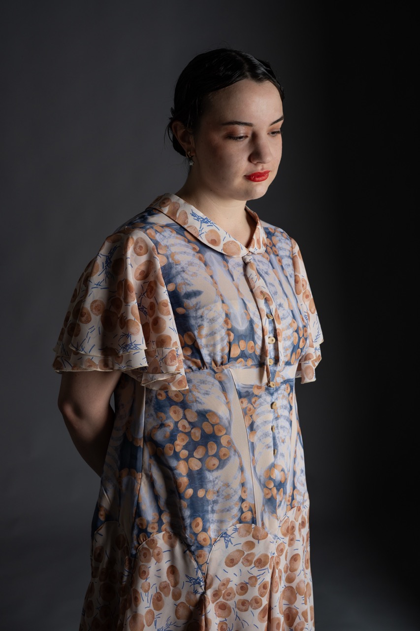 A young woman with dark hair wearing a light-coloured dress featuring a floral and polka dot pattern. She stands with her arms behind her back, looking down, against a dark, neutral background that highlights her silhouette.