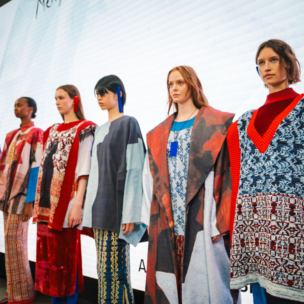 Five models on catwalk wearing layered garments with geometric patterns, red accents, and mixed prints in blue, white, and earth tones.