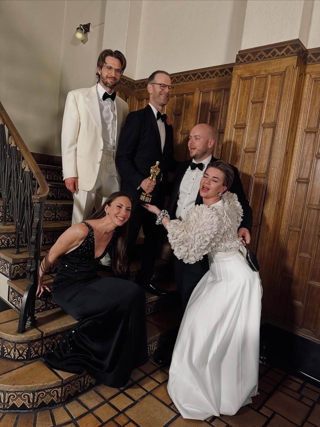 Five people in formal evening wear posing on ornate wooden staircase. Man in white dinner jacket, others in black formal attire and white gown.