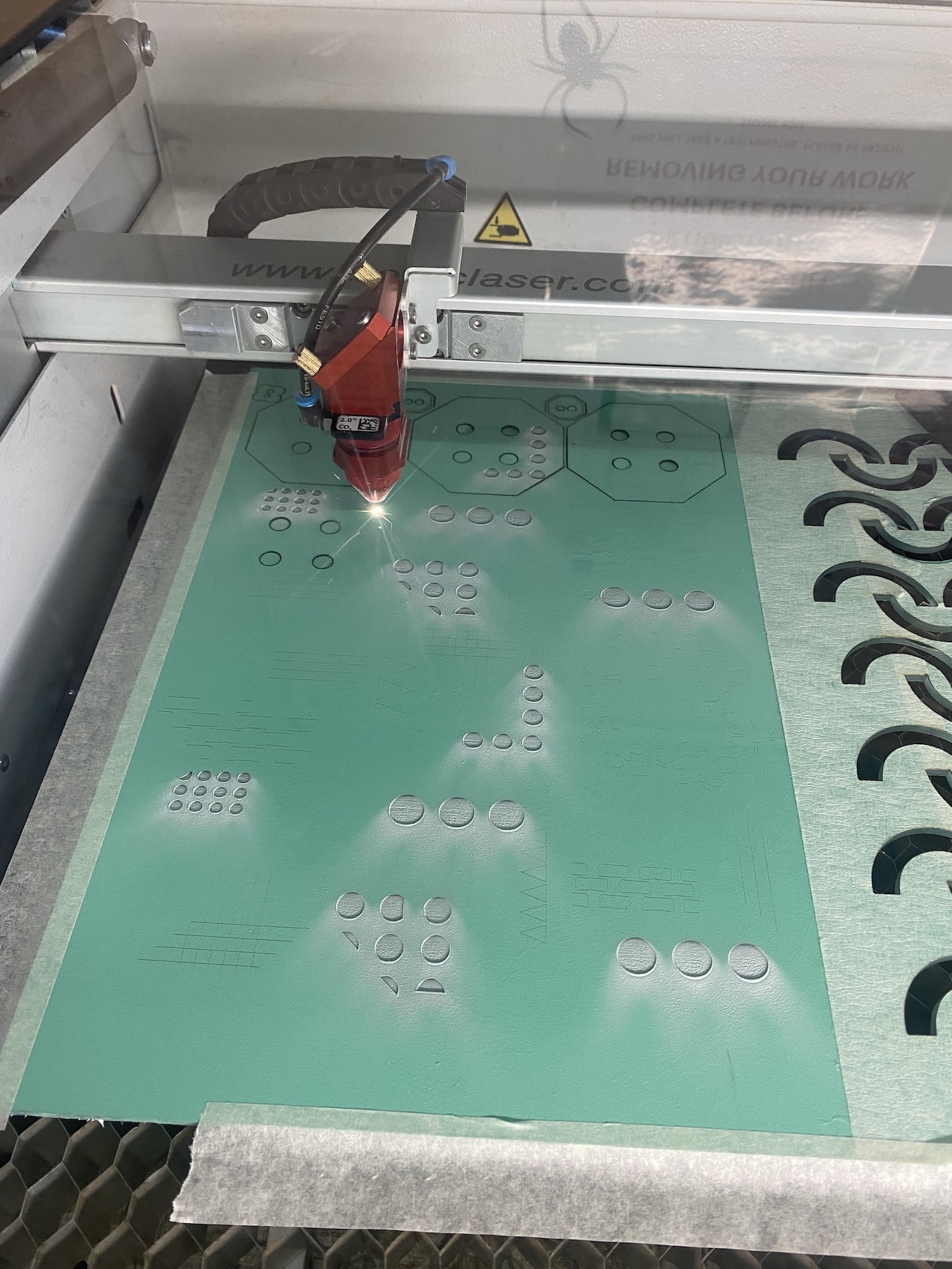 Laser cutting machine with red cutting head operating on green acrylic sheet with circular and rectangular cutout patterns.