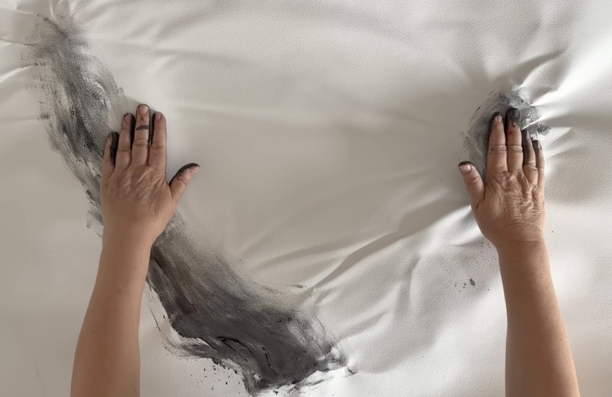 Two hands apply black charcoal to a white canvas, creating soft smudges and texture in the material. The canvas is slightly wrinkled, and the hands are positioned on either side of the area being worked on.