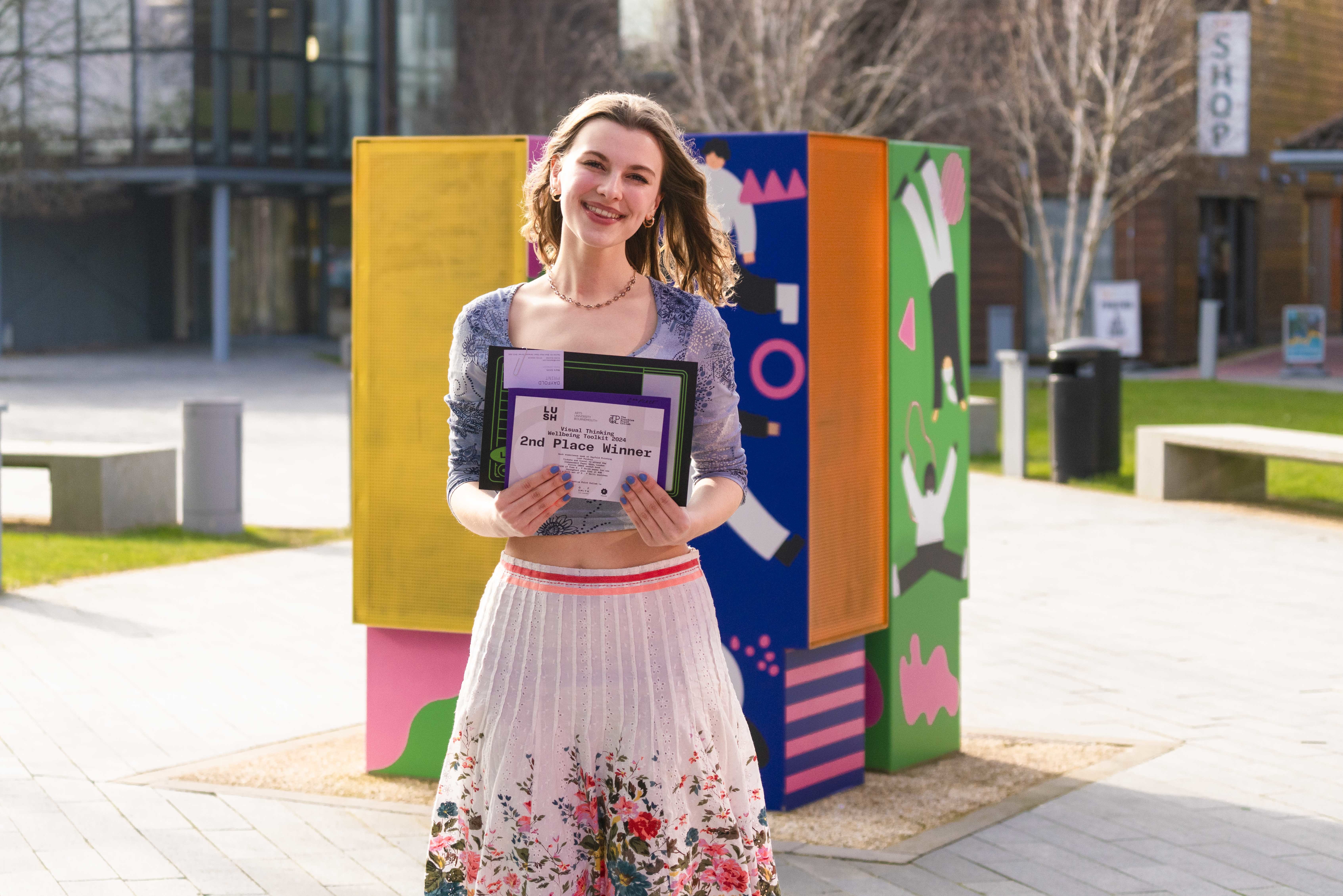 Lauren Gardner stood outside on-campus holding a 2nd place certificate, wearing floral clothing.