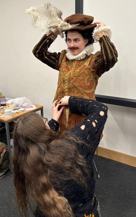 Student being dressed in 17th century-style male costume, brown themed, including a hat with a feather.