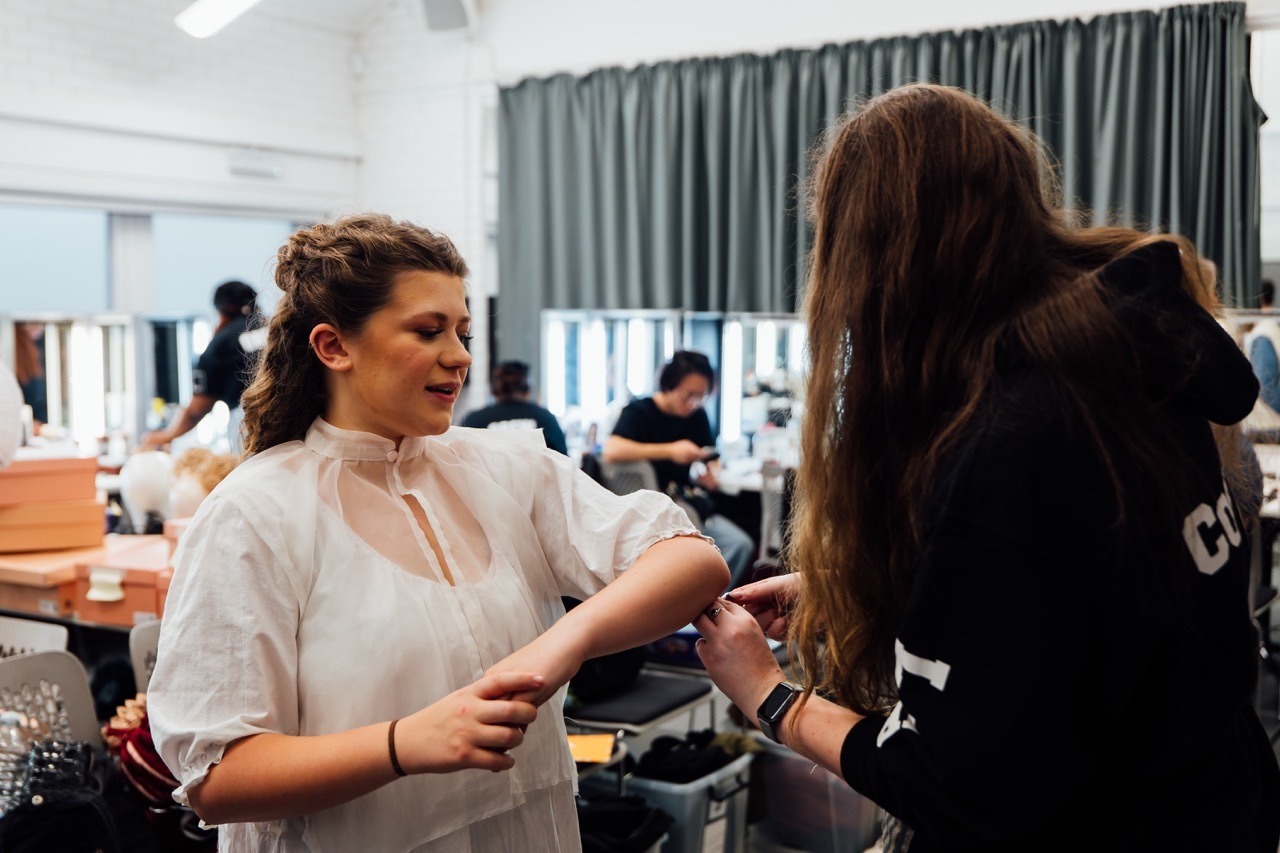 Woman in white blouse having hair styled by hairdresser with long brown hair in salon with green curtains and other clients.