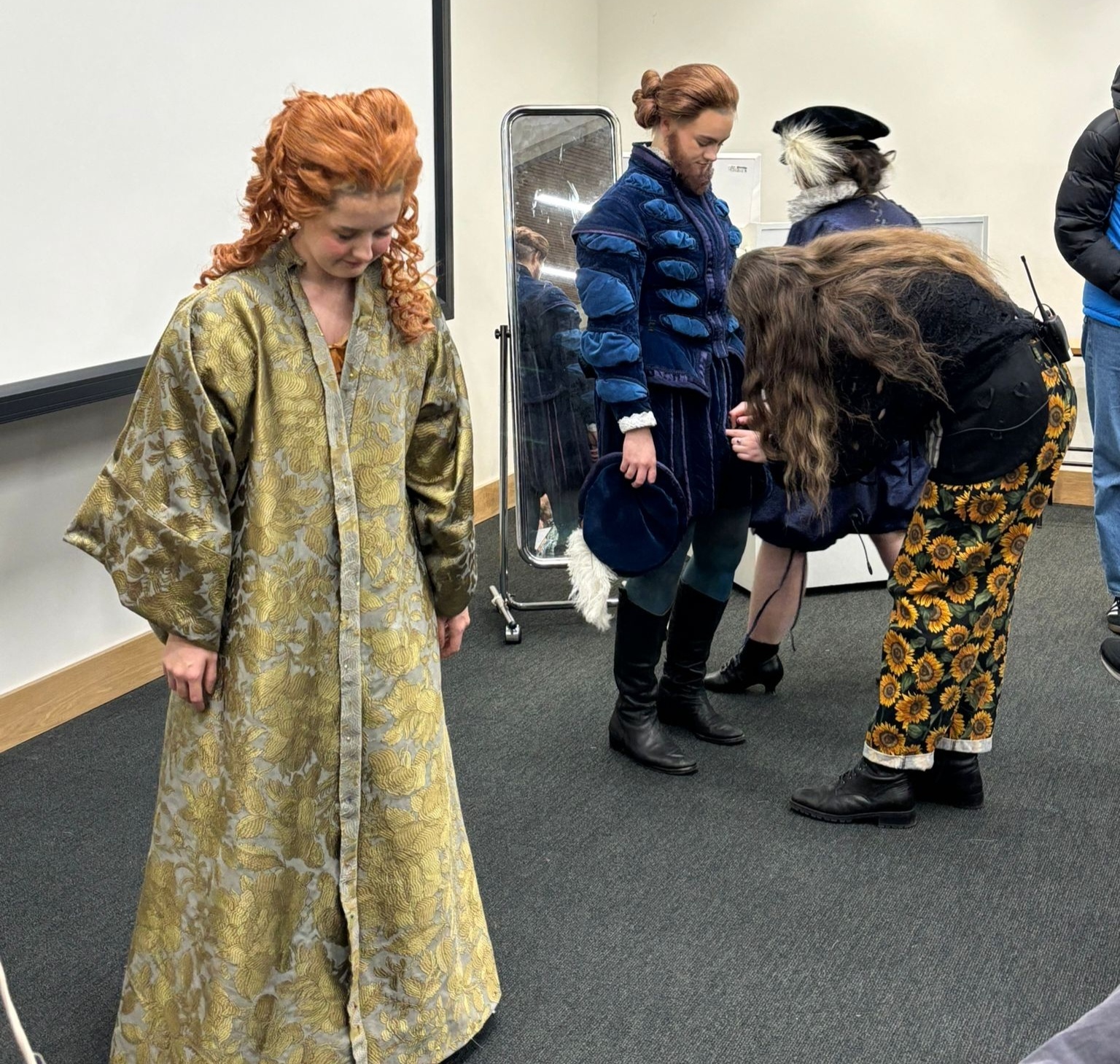 Woman in elaborate red curly wig and green patterned robe stands beside mirror whilst others in blue coats assist with costume fitting backstage.