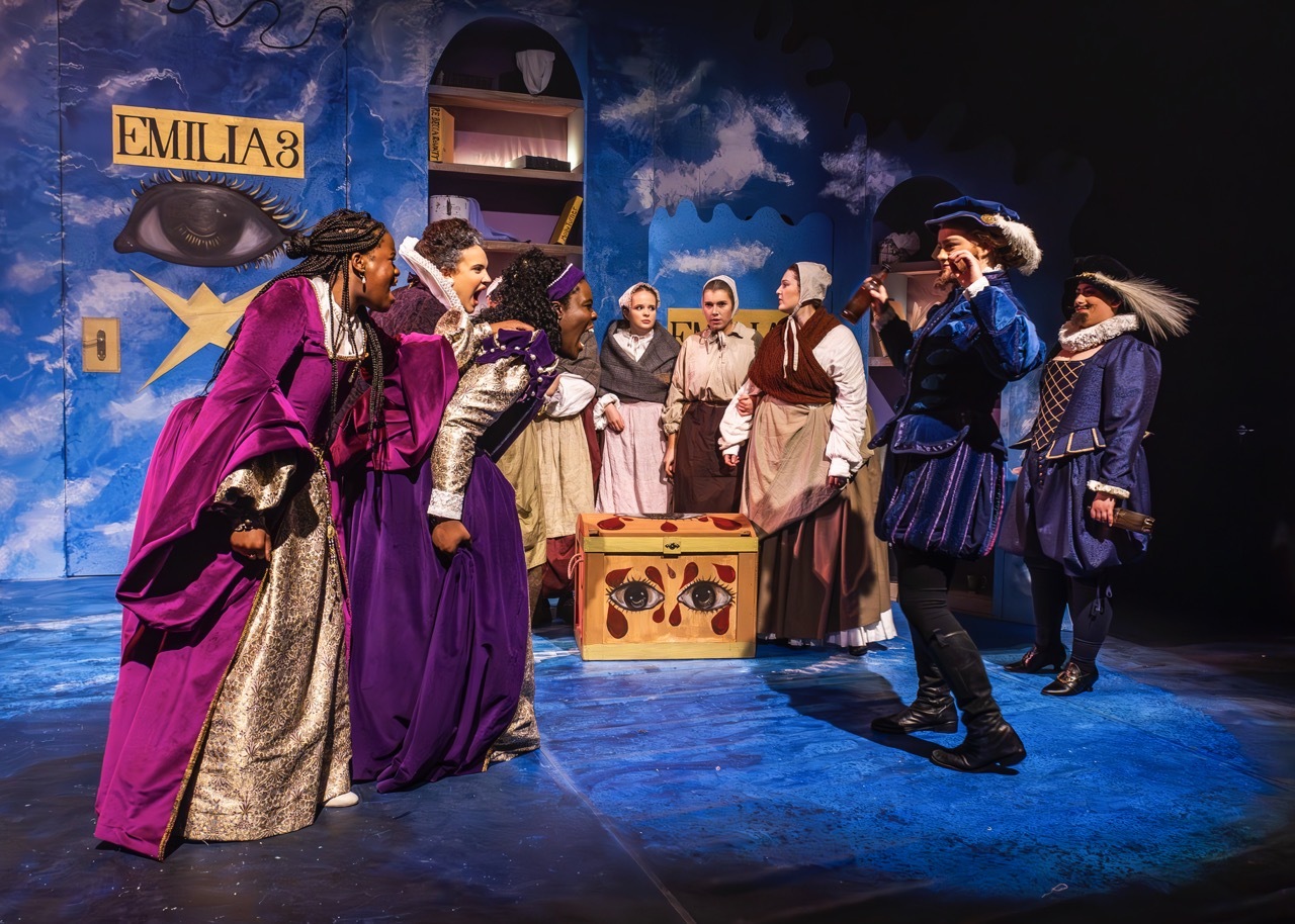 Theatre performers in period costumes on blue-lit stage with painted backdrop featuring ship and "EMILIA" sign.