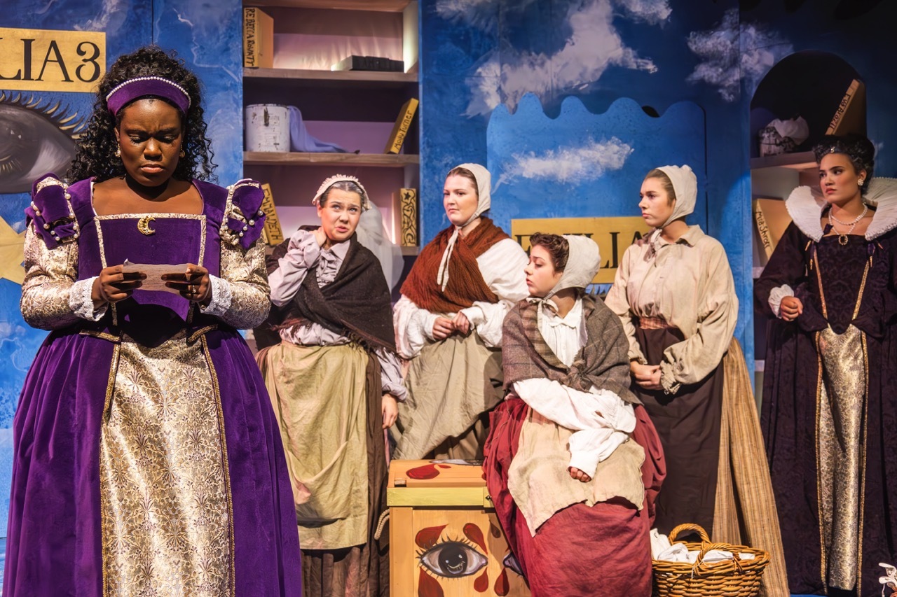 Theatre performers in period costumes on stage with blue sky backdrop, wooden shelving, and props including baskets and books.