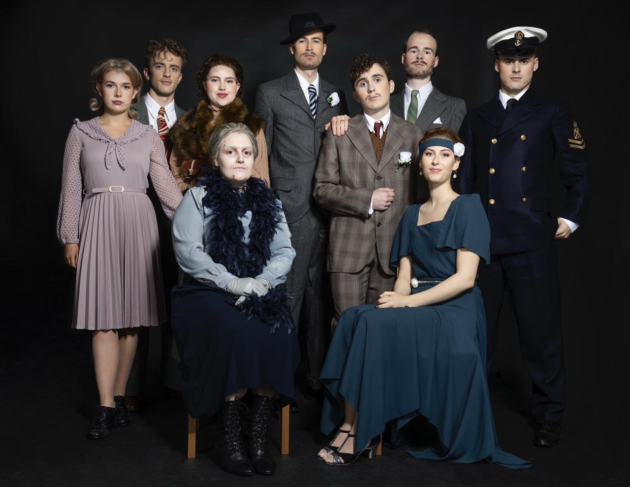 Cast of nine actors in period costume posing for group portrait. Woman in mauve dress, elderly woman seated in navy, man in naval uniform, others in 1940s attire.
