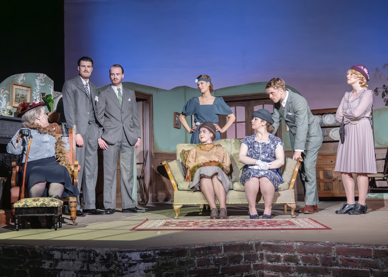 Eight actors on stage in period costume performing in living room set with green walls, furniture, and patterned rug against blue backdrop.