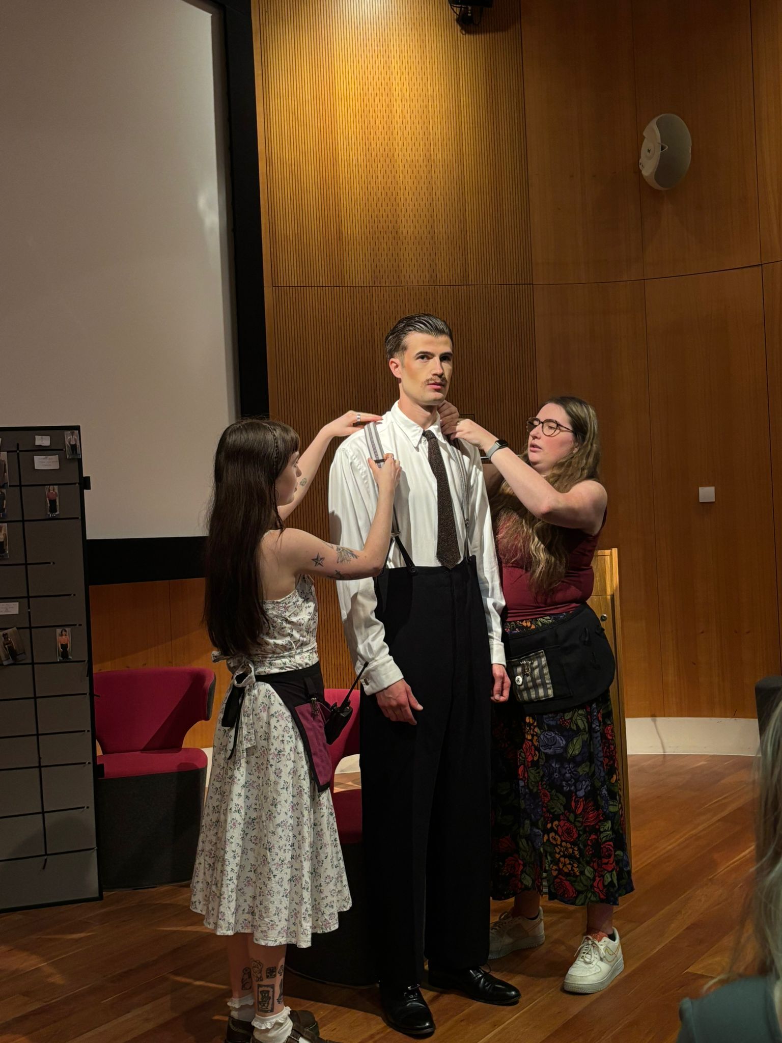 Man in white shirt and black trousers being fitted with red braces by two women on wooden stage with brick wall backdrop.