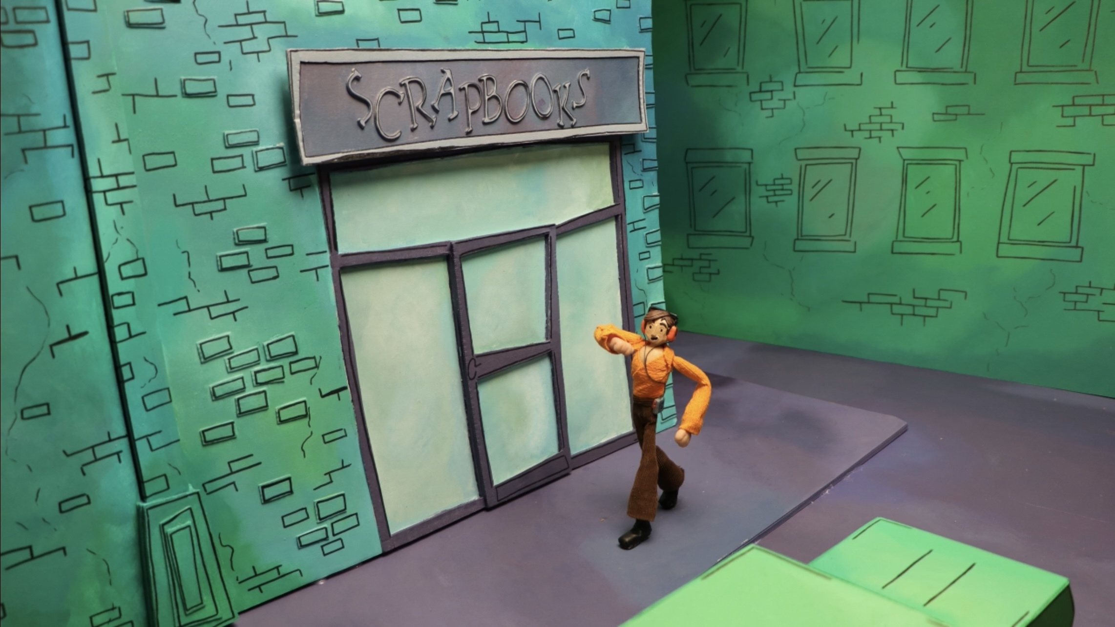 Animation still of an orange character walking down a street with green buildings, outside a shop with windows named 'scrapbook'.