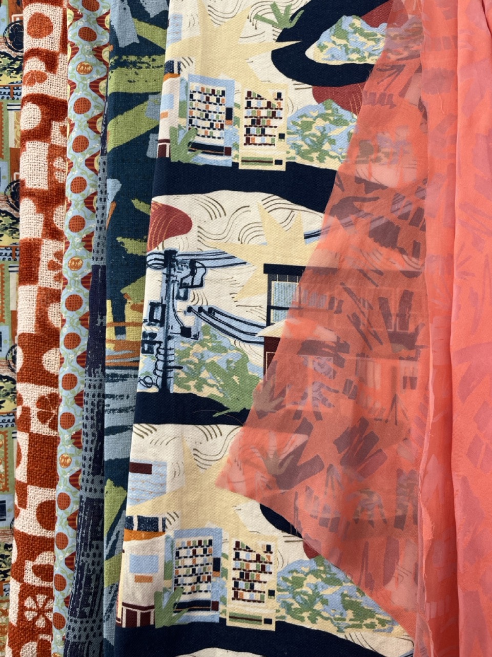 A close-up view of several fabrics displayed side by side. The fabrics feature a variety of patterns, including geometric shapes in red and blue, a print with stylised buildings amid trees, and a sheer coral material.