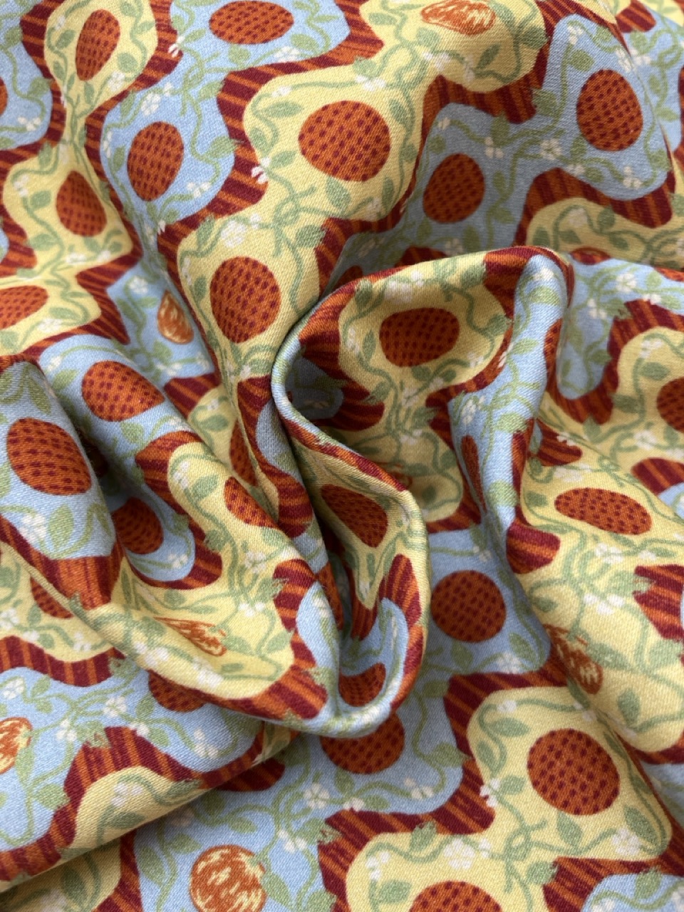 A close-up of fabric featuring a repeating pattern of large red strawberries interspersed with blue flowers on a light yellow background. The fabric is draped, creating folds and shadows that highlight the design.