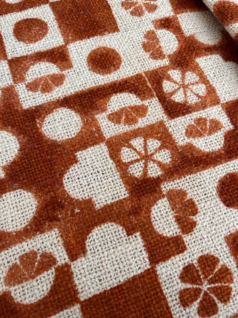A close-up view of a patterned fabric featuring a mix of orange and beige designs, including circles, floral shapes, and geometric squares. The textures of the weave are visible, highlighting the intricate patterns on the surface.