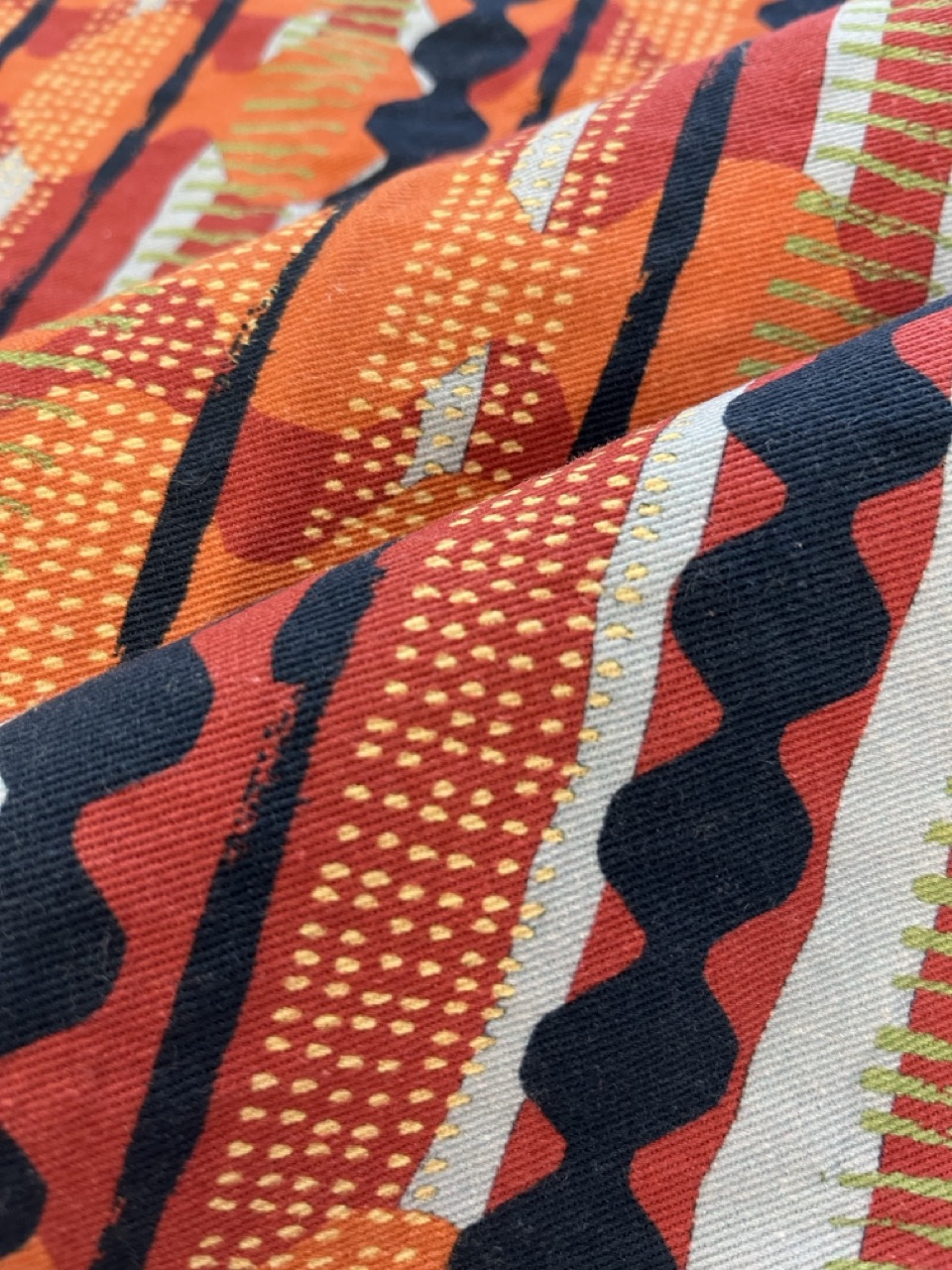 A close-up of fabric displaying a vibrant pattern with alternating stripes in red, orange, and navy blue. The design includes various geometric shapes and textures, such as dots and zigzag lines, creating a visually dynamic effect.