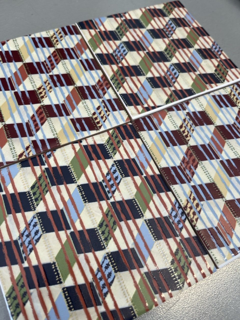 Four square tiles arranged in a grid pattern, each displaying a distinctive geometric design. The designs feature stripes and coloured blocks in tones of red, blue, green, and cream against a patterned background.