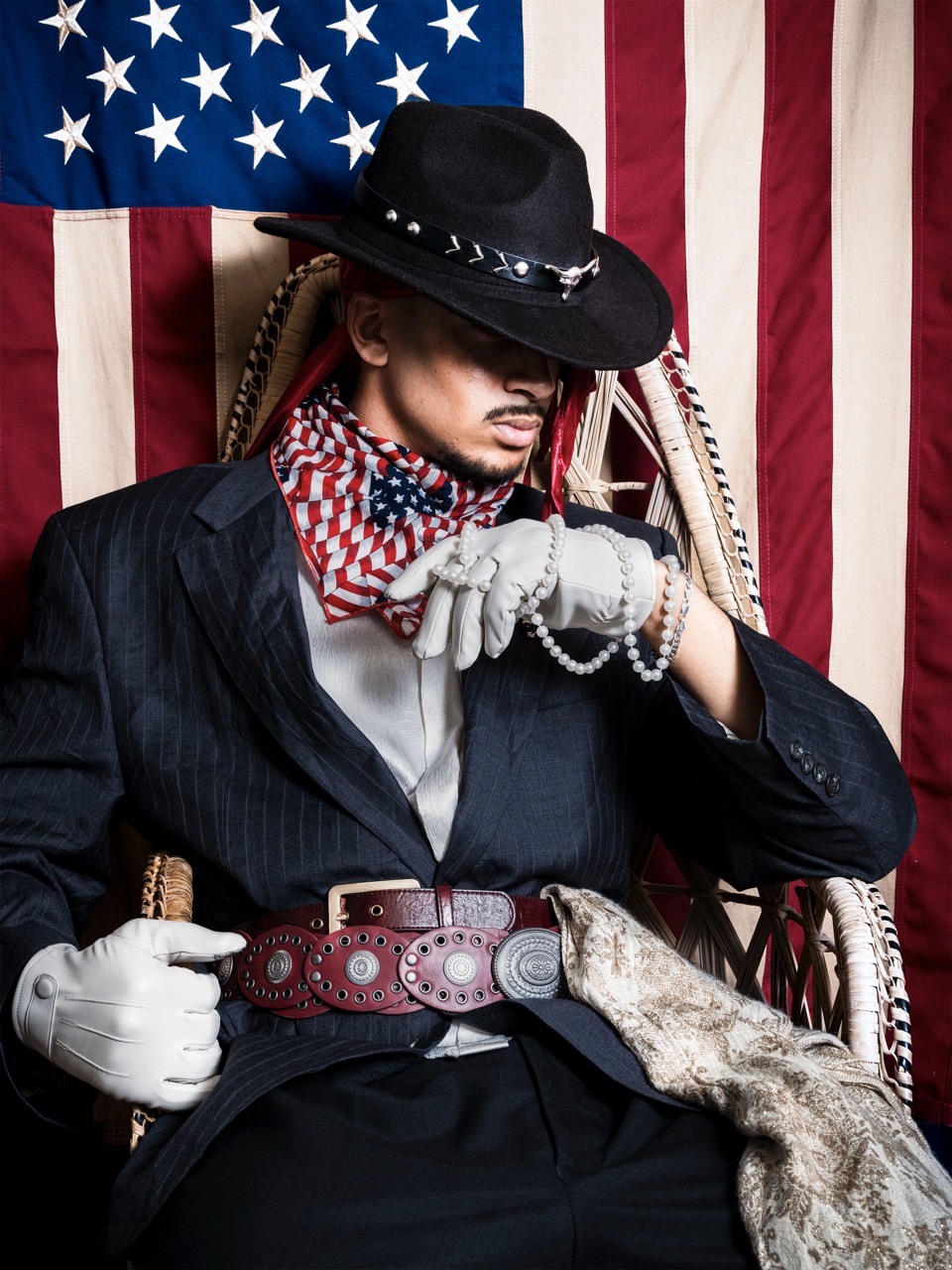 A man wearing a black hat, a striped jacket, and a red and white bandana poses with a silver chain in front of an American flag. He is seated on a wicker chair, and a patterned fabric drapes over his lap.