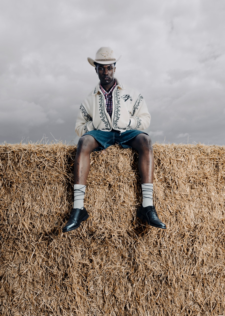 A man sits on a hay bale wearing a white jacket with black embroidery, a checked shirt, and a cowboy hat. He has blue shorts and white socks, with black shoes. The background features an overcast sky, creating a rustic outdoor setting.