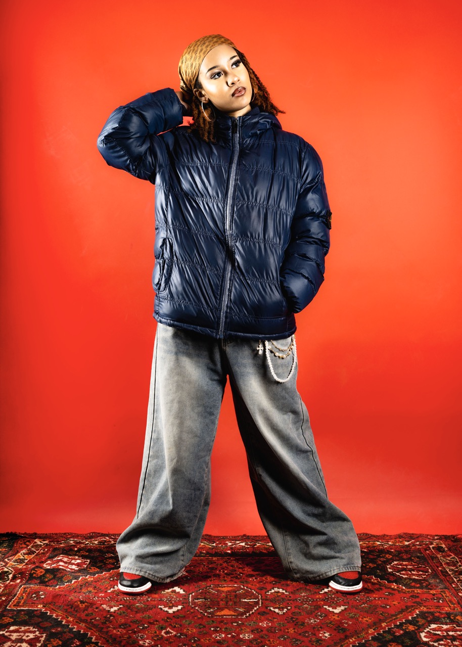 A young person stands against a red background wearing a navy blue puffer jacket and grey wide-leg trousers. They have shoulder-length brown hair with lighter highlights and are positioned on a patterned rug.