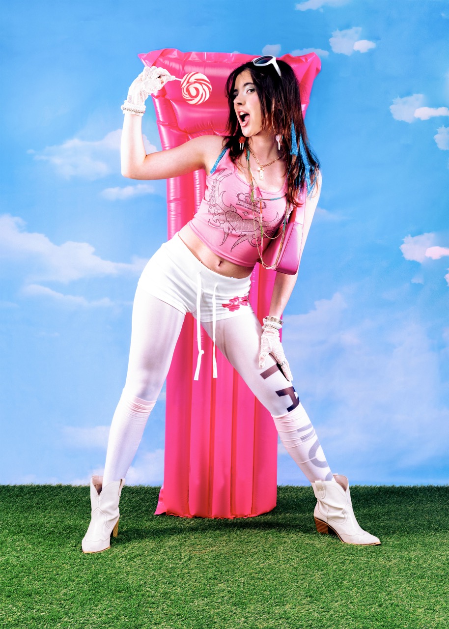 A woman in a pink crop top and white leggings stands confidently, posing with her hands on her hips. She wears white heeled boots and oversized sunglasses, and is in front of a bright blue sky backdrop with fluffy clouds and a pink inflatable float.