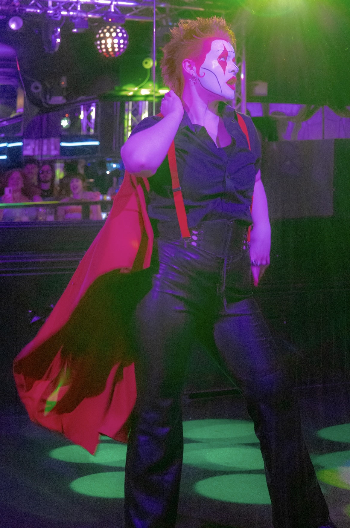 Drag performer posing on stage bathed in purple, green and magenta lighting with stage lights visible behind.