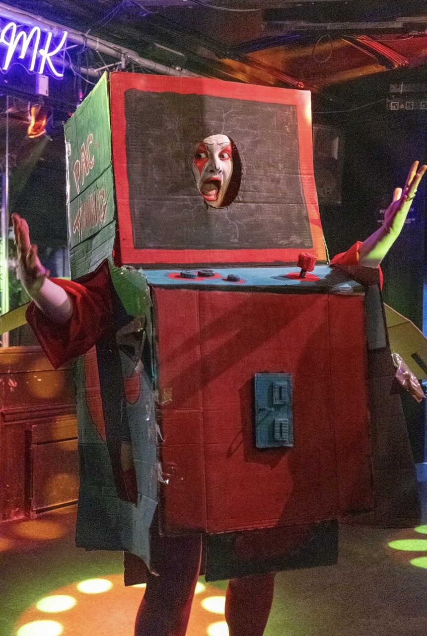 Person in red rectangular arcade costume with face visible through screen opening, green accents on sides, neon lighting in background.