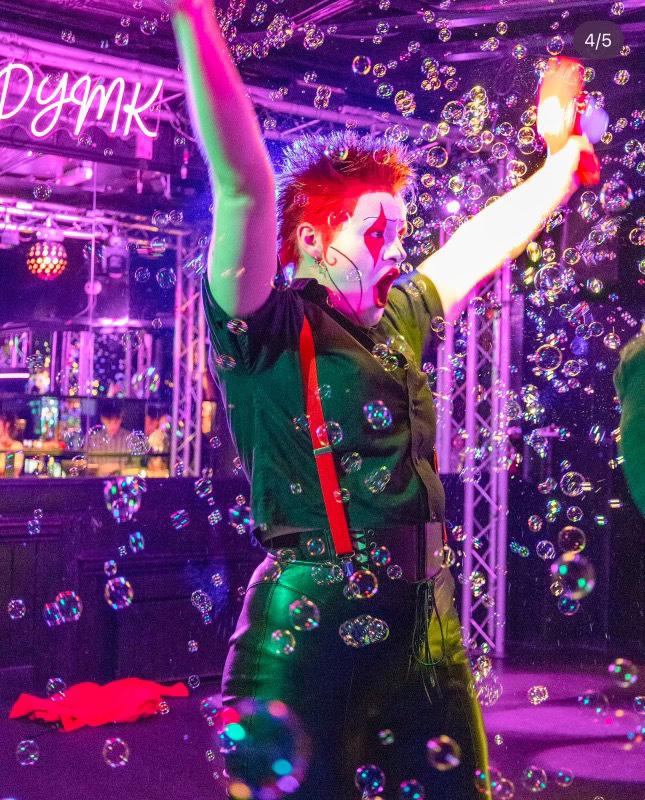 Person with arms raised in nightclub with purple neon lighting, soap bubbles floating throughout scene, and bar visible in background.