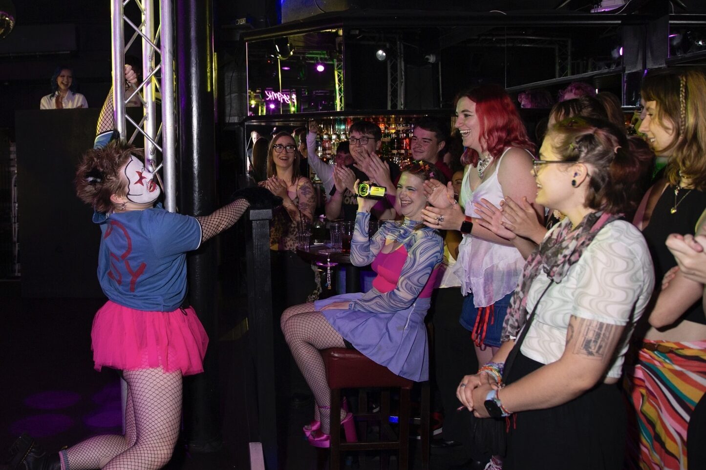 Performer in blue top and pink tutu interacting with seated audience in dimly lit venue with metal framework and purple lighting.