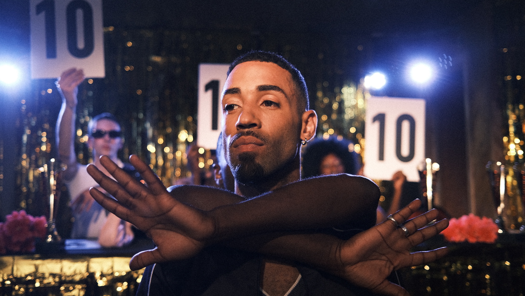 Close-up of an actor from Realness with a Twist, posing with their arms crossed and palms facing forwards. Other people are holding cards with the number 10 on them in the background.