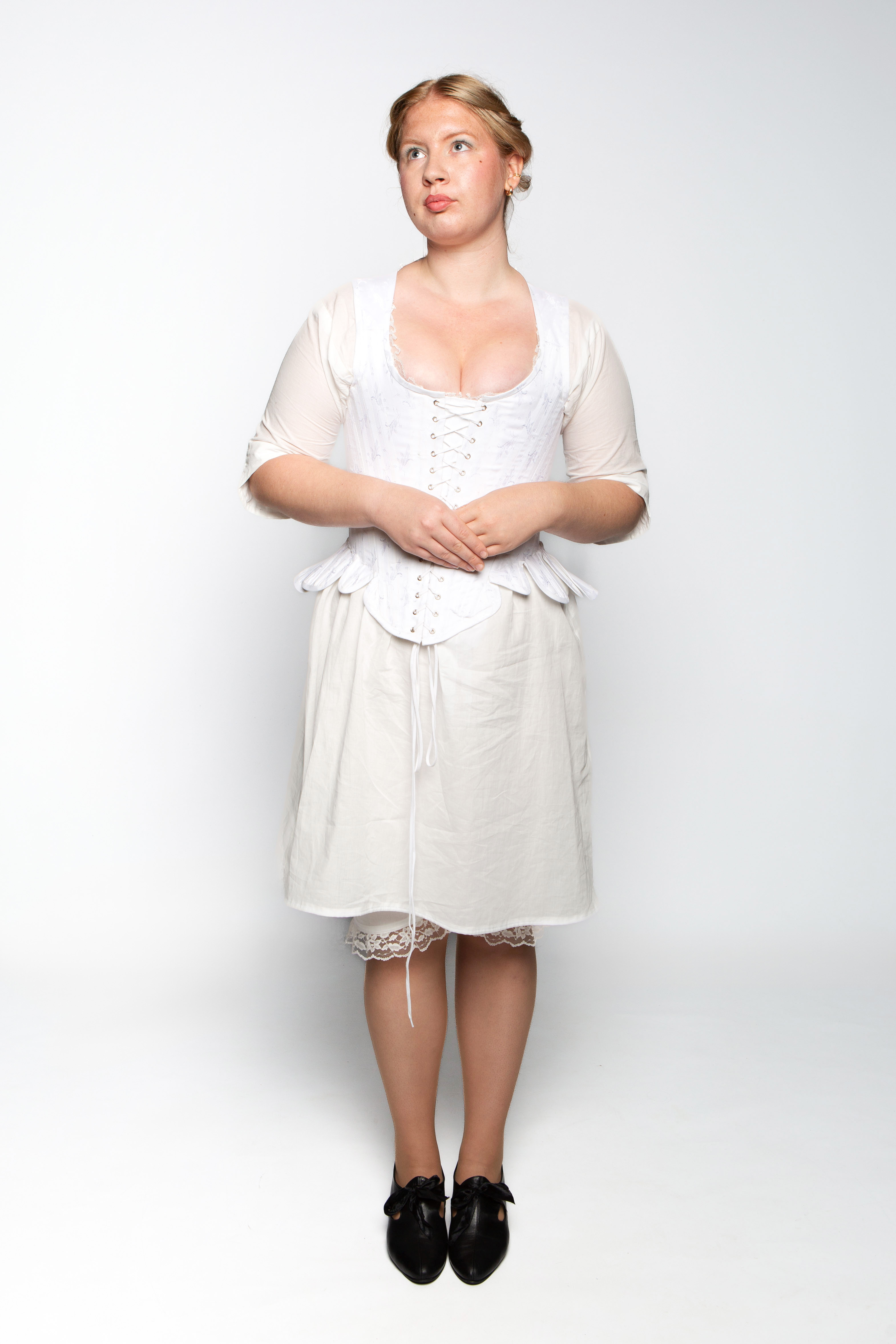 A woman stands upright, wearing a white historical dress with a fitted bodice and a flowing skirt, paired with black shoes. Her hair is styled in a simple updo, and she is positioned against a plain white background.