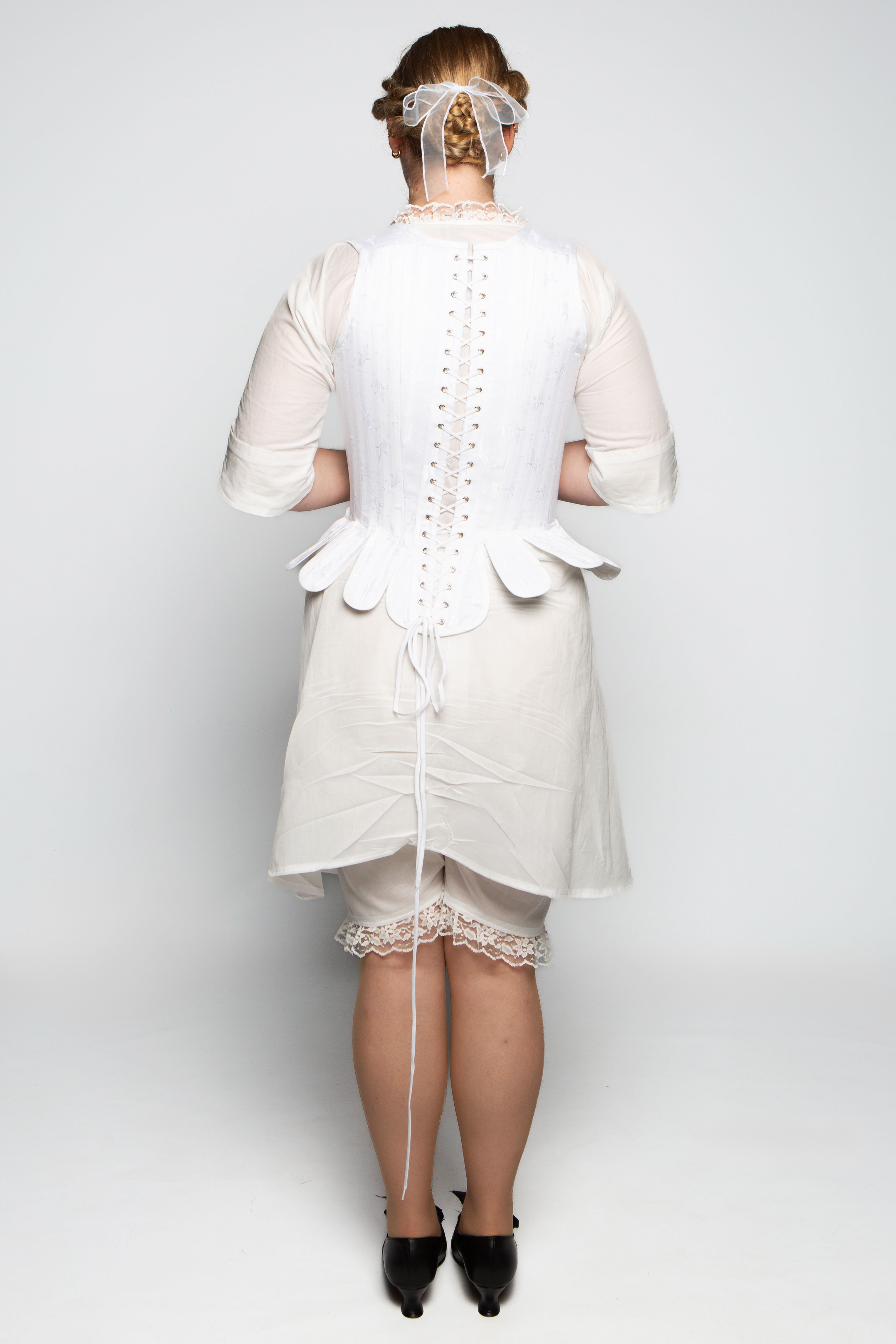 A woman wearing a vintage-style dress with a fitted bodice and tied elements at the back. The dress is off-white, with short sleeves, and has a lace trim visible underneath. Her hair is styled with a decorative headpiece.