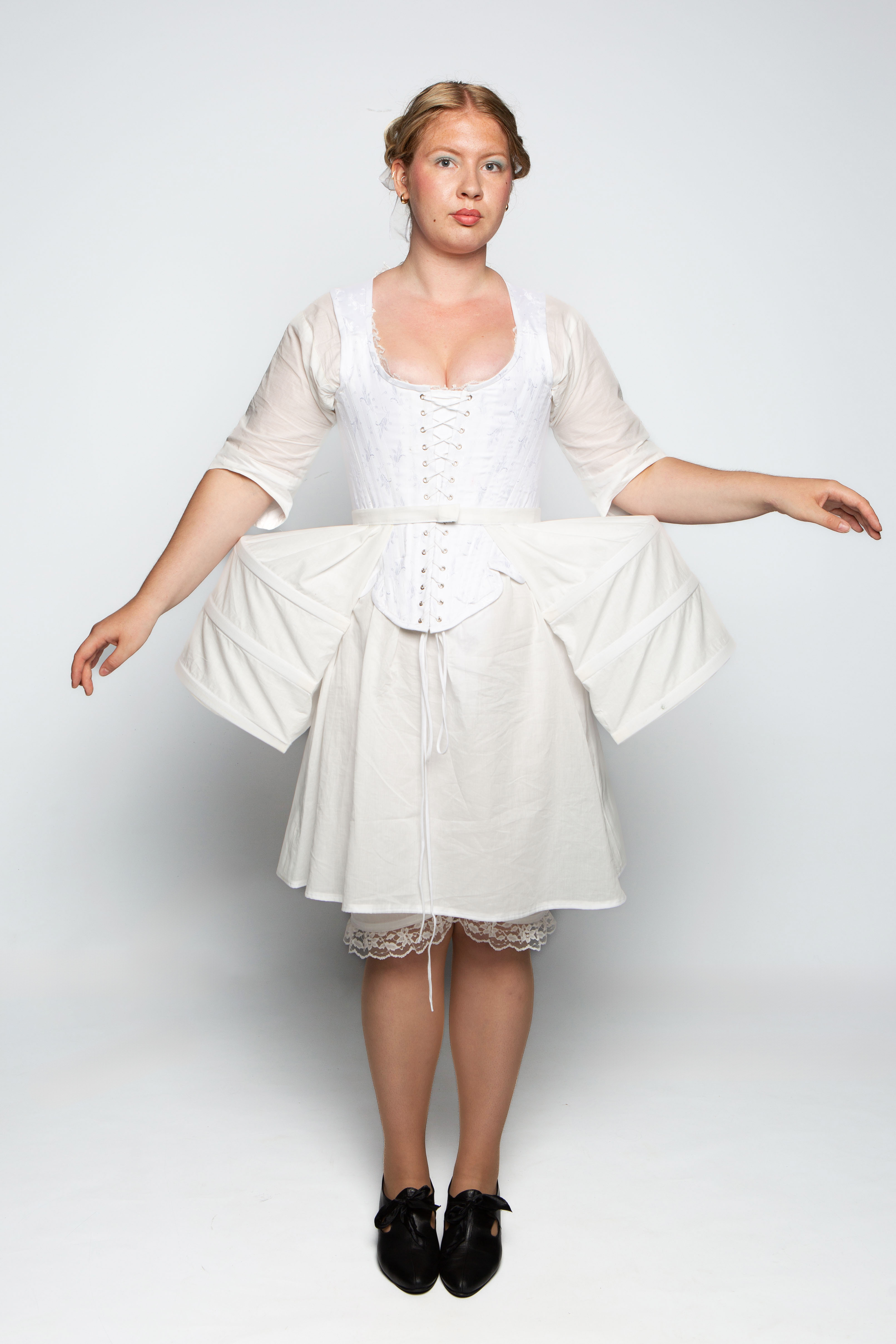 A woman stands wearing a white dress with puffed sleeves and a corset top. The dress features a fitted bodice and a full, skirt-like bottom with exaggerated side panniers. She has her arms extended outwards and is barefoot.