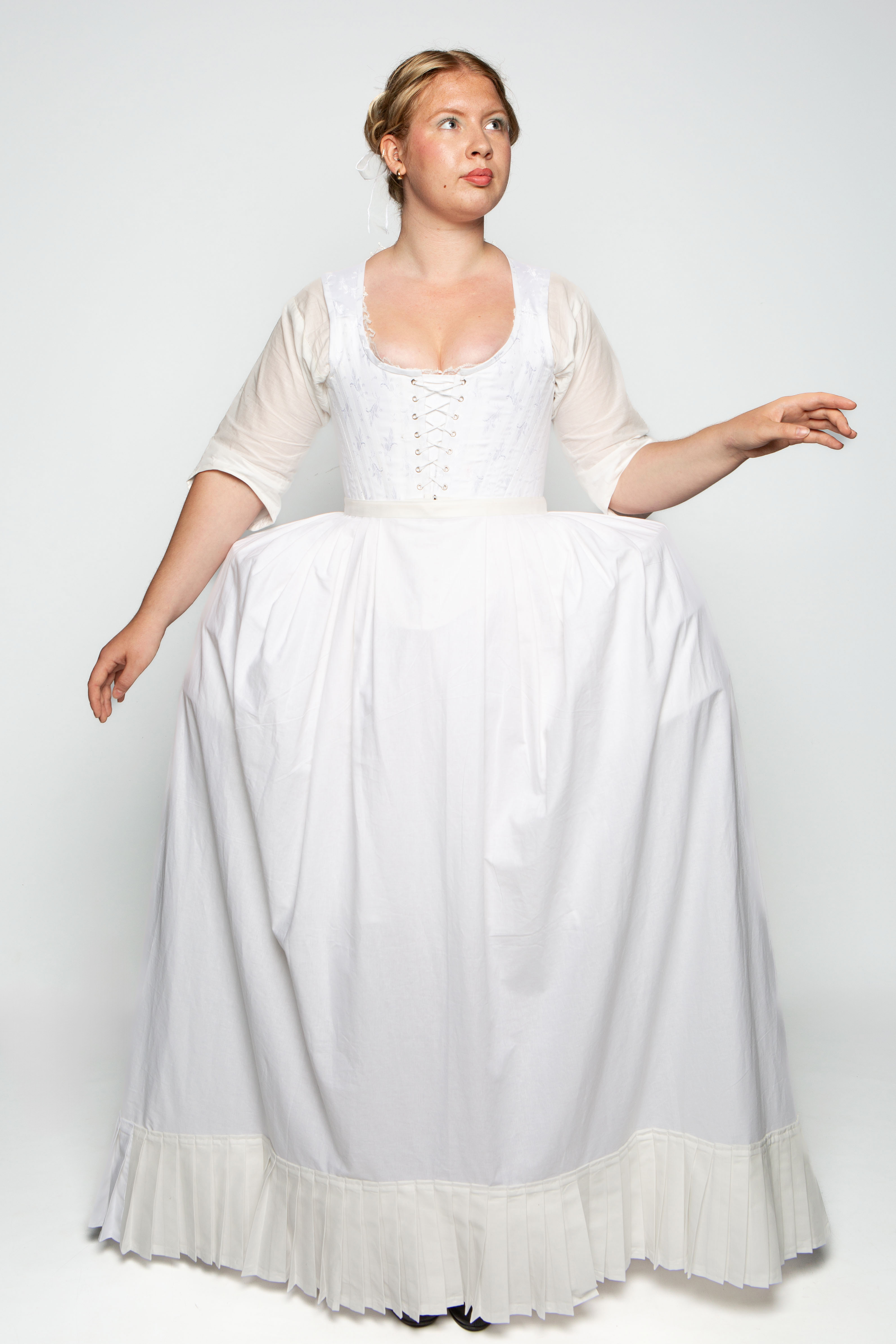 A woman wearing a white historical dress with a fitted bodice and full skirt. The dress features a lace-up front and long, sheer sleeves, with a ruffled hemline. She stands against a plain, light background.
