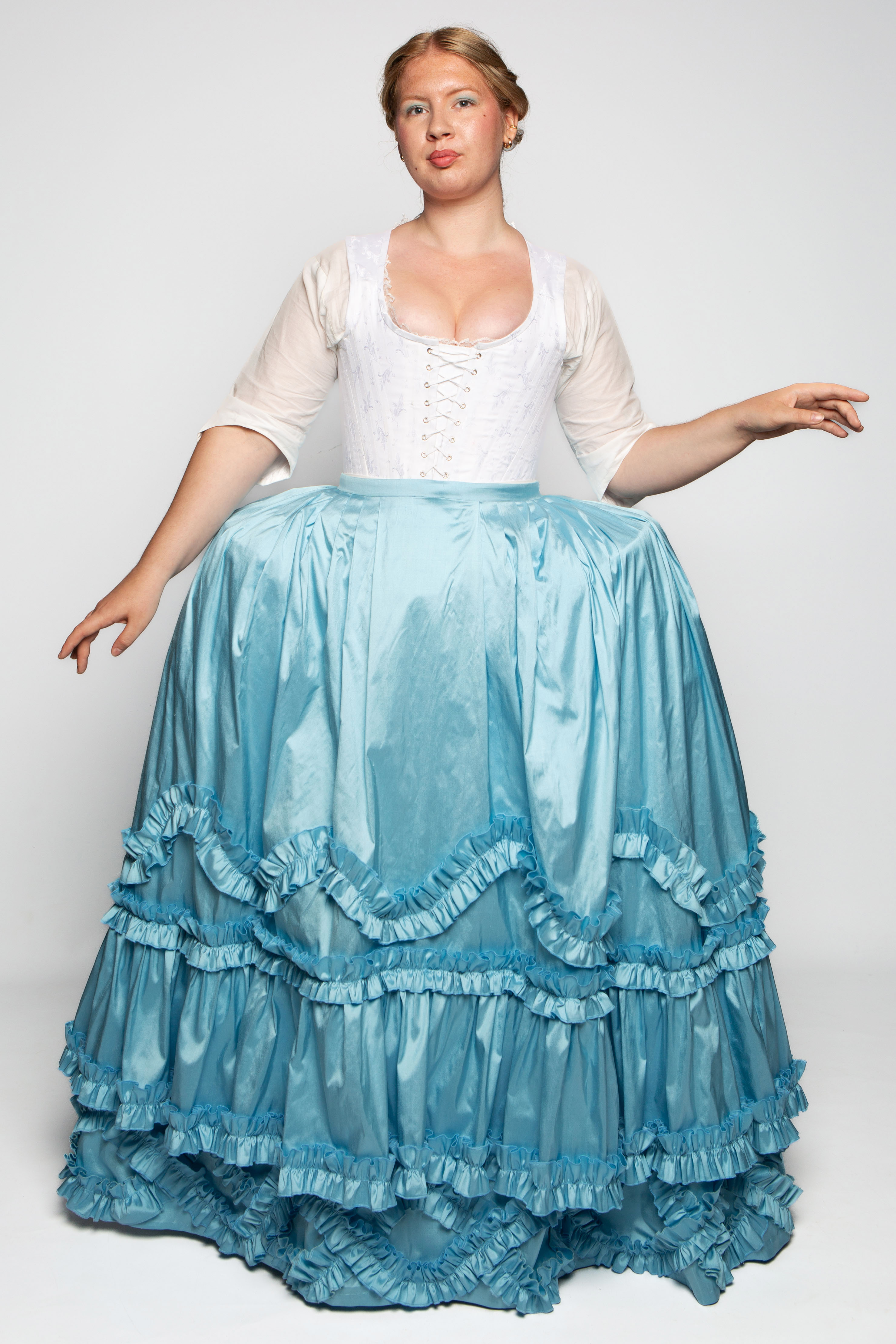 A woman wearing a white bodice and a long blue satin skirt with ruffles stands against a plain background. The skirt is full and has multiple layers, featuring gathered fabric along the edges.