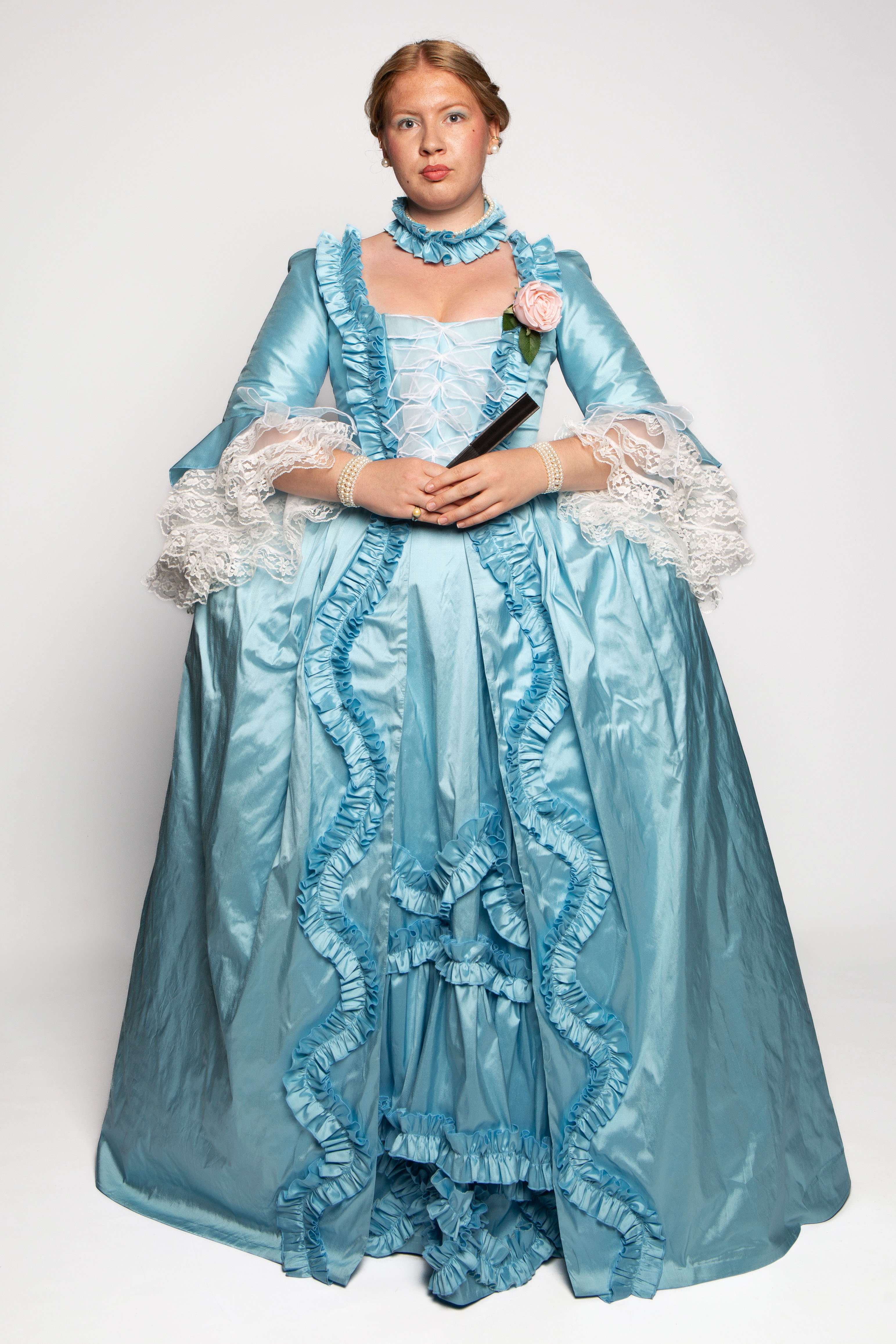 A young woman in an elaborate light blue gown with ruffled details, lace sleeves, and a matching necklace holds a small bouquet and stands against a plain backdrop. The gown features a voluminous skirt and is styled in a historical fashion.