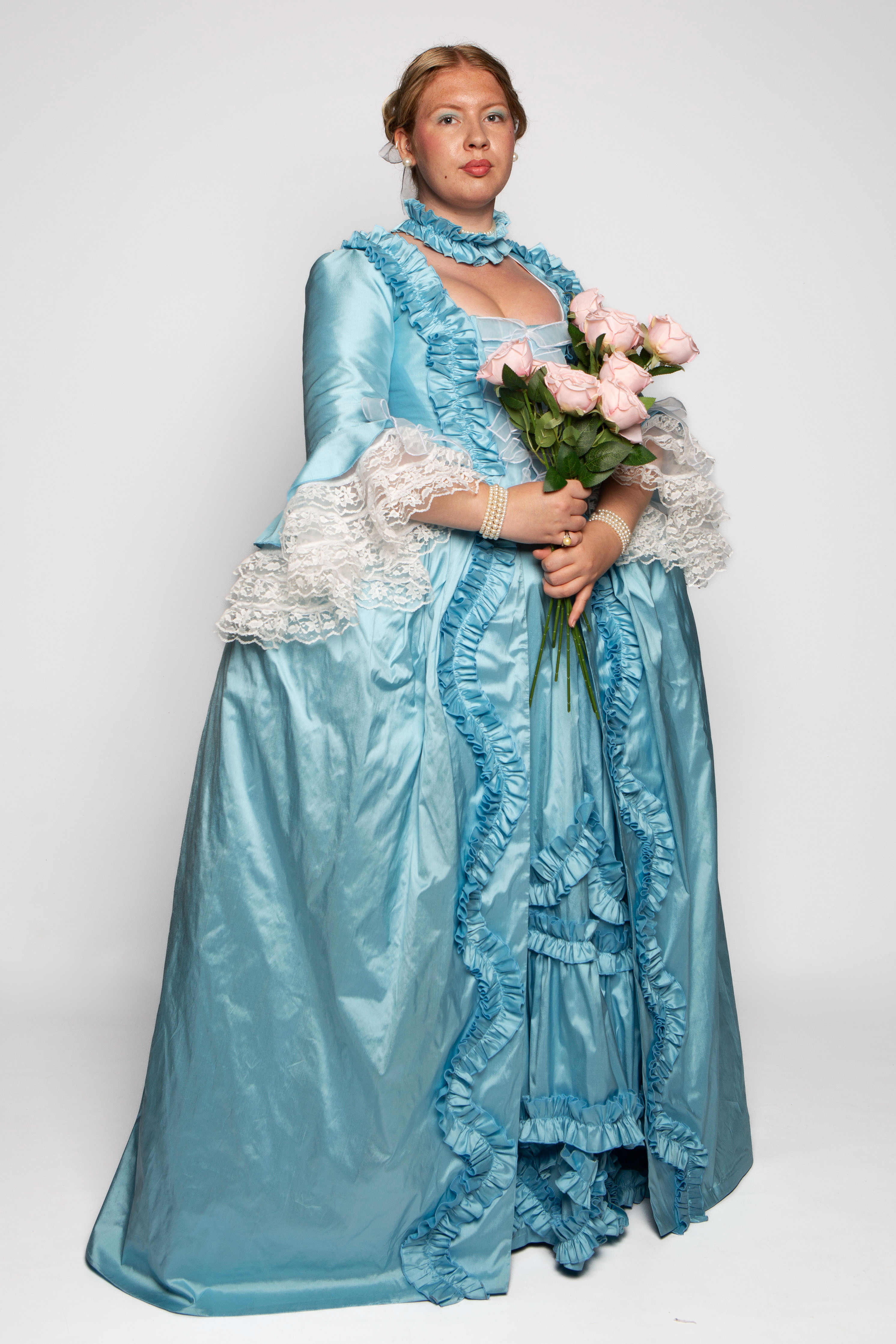A woman wearing an elaborate light blue dress with lace details stands holding a bouquet of pink roses. The dress features ruffles and a collar, with she has styled hair and light makeup. The background is a plain, light colour.