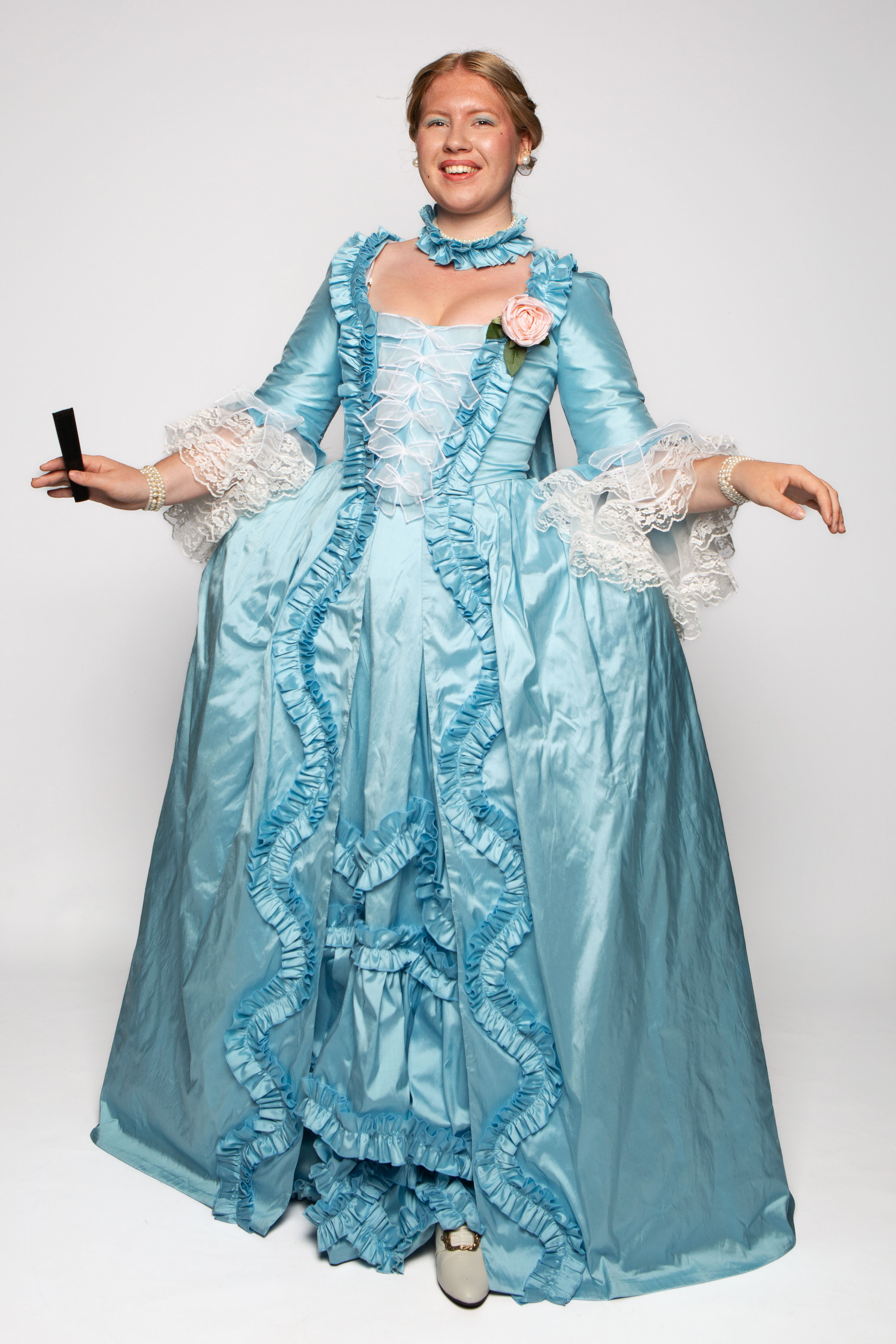 A woman wearing a light blue gown with ruffled details and a floral decoration on the chest stands with her arms outstretched. The gown features lace accents and a full skirt, against a plain white backdrop.