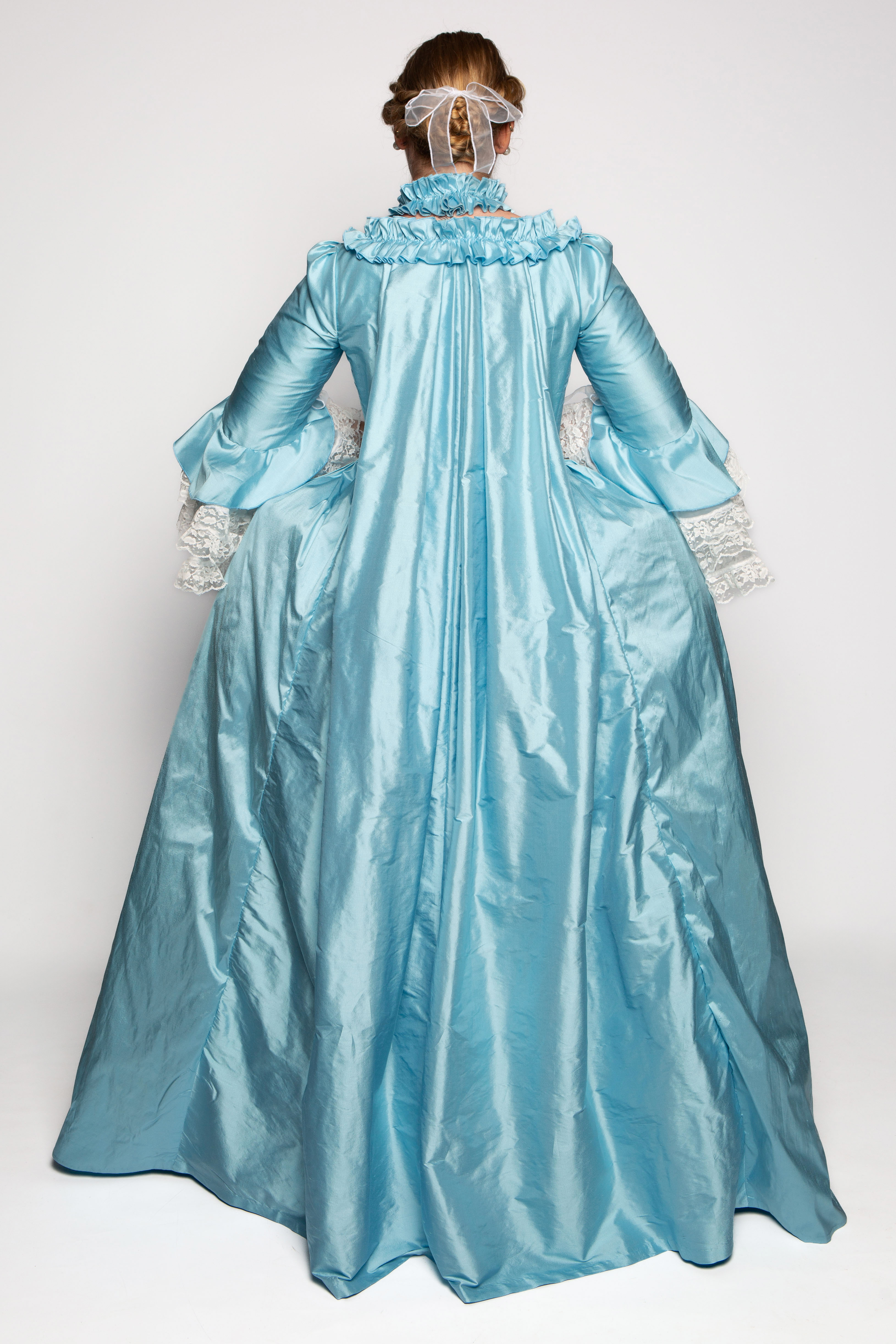 A woman with her back to the camera wears a long, flowing blue gown. The dress features a high neck with ruffles and wide sleeves, along with intricate white lace cuffs. The hair is styled in an updo, adorned with a decorative headpiece.