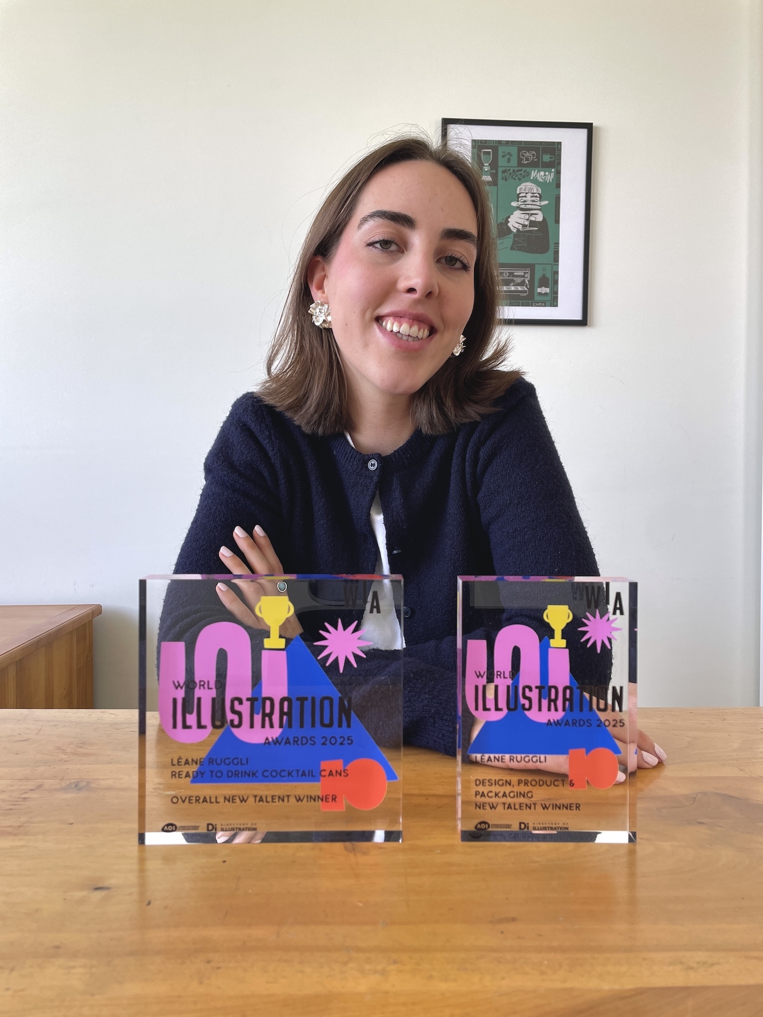L Ruggli smiling behind wooden table with two clear acrylic awards featuring pink and blue text. Framed artwork on white wall.