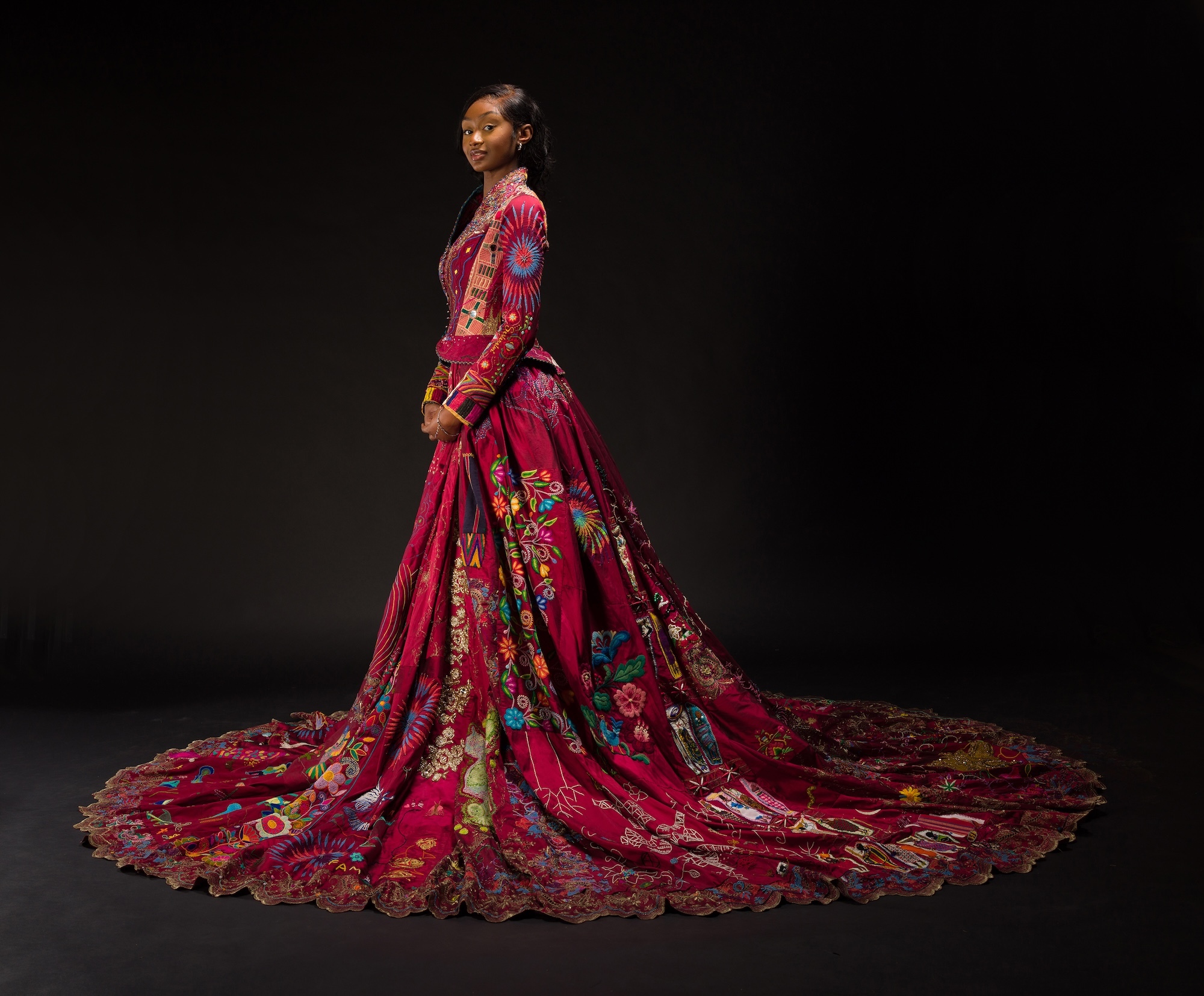 Woman in profile wearing elaborate burgundy gown with floral patterns and dramatic floor-length train against black background.