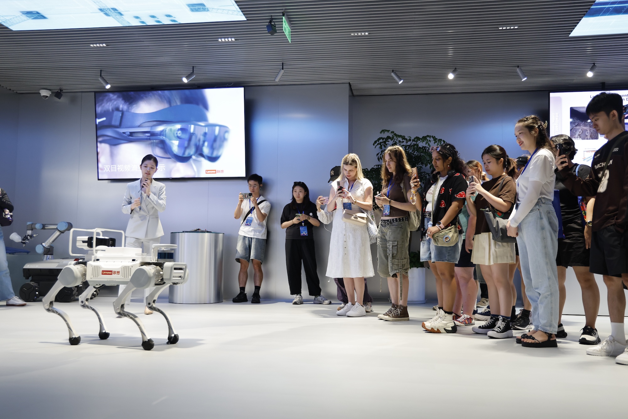 A white four-legged robot dog stands in a modern showroom, with a presenter and a group of visitors watching beside a large screen in the background.