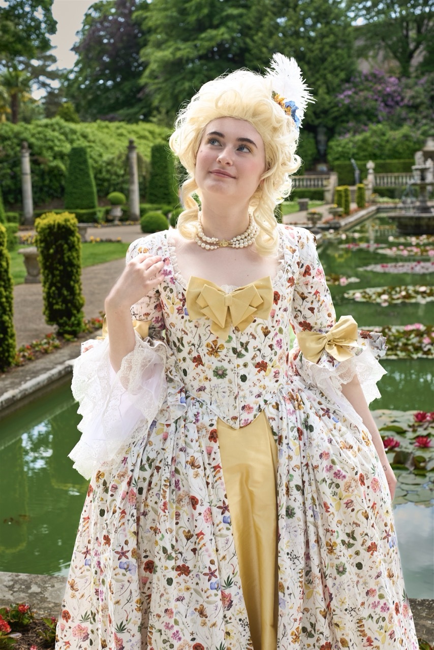 Woman in 18th-century costume with blonde powdered wig, floral gown, and pearl necklace standing in formal garden with pond.