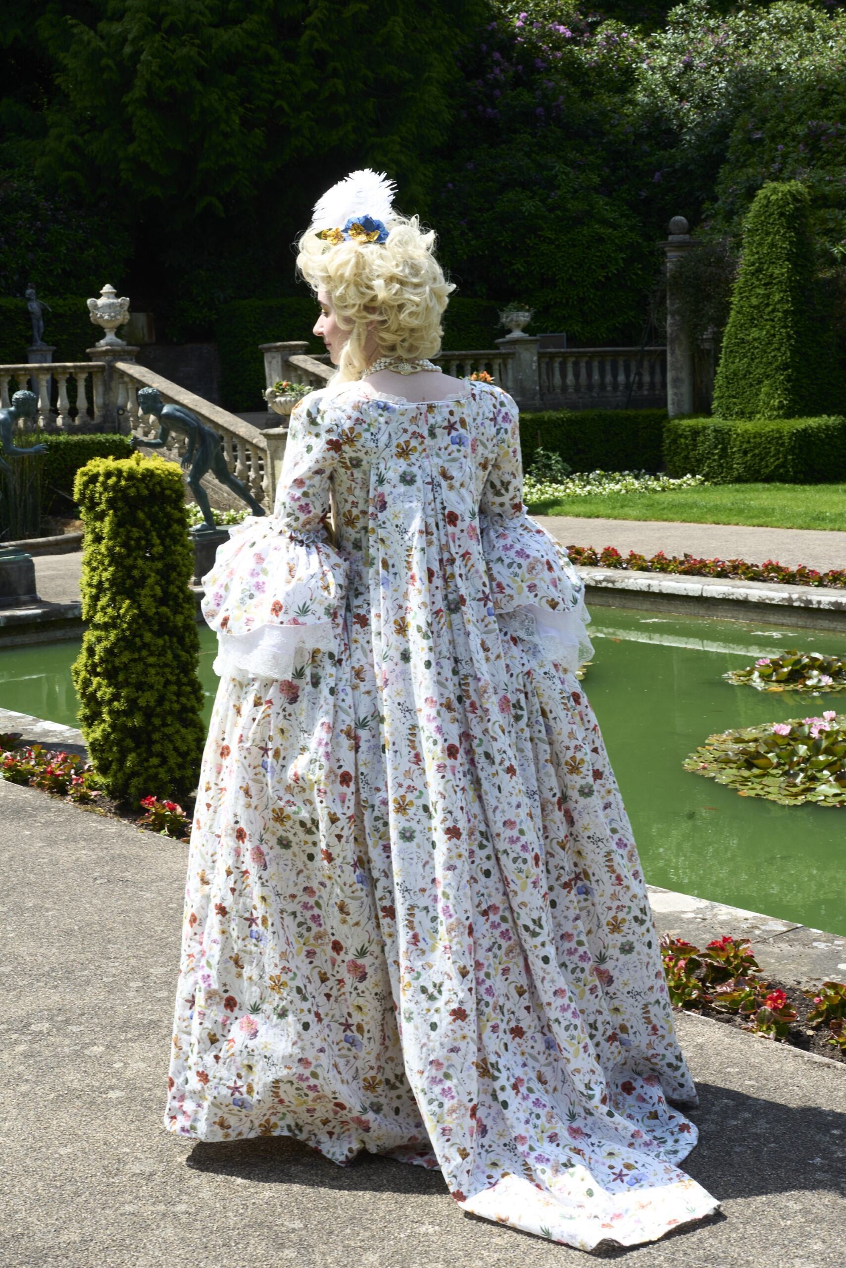 Person in 18th-century white floral gown with powdered wig stands beside ornamental pond in formal garden with stone balustrade.