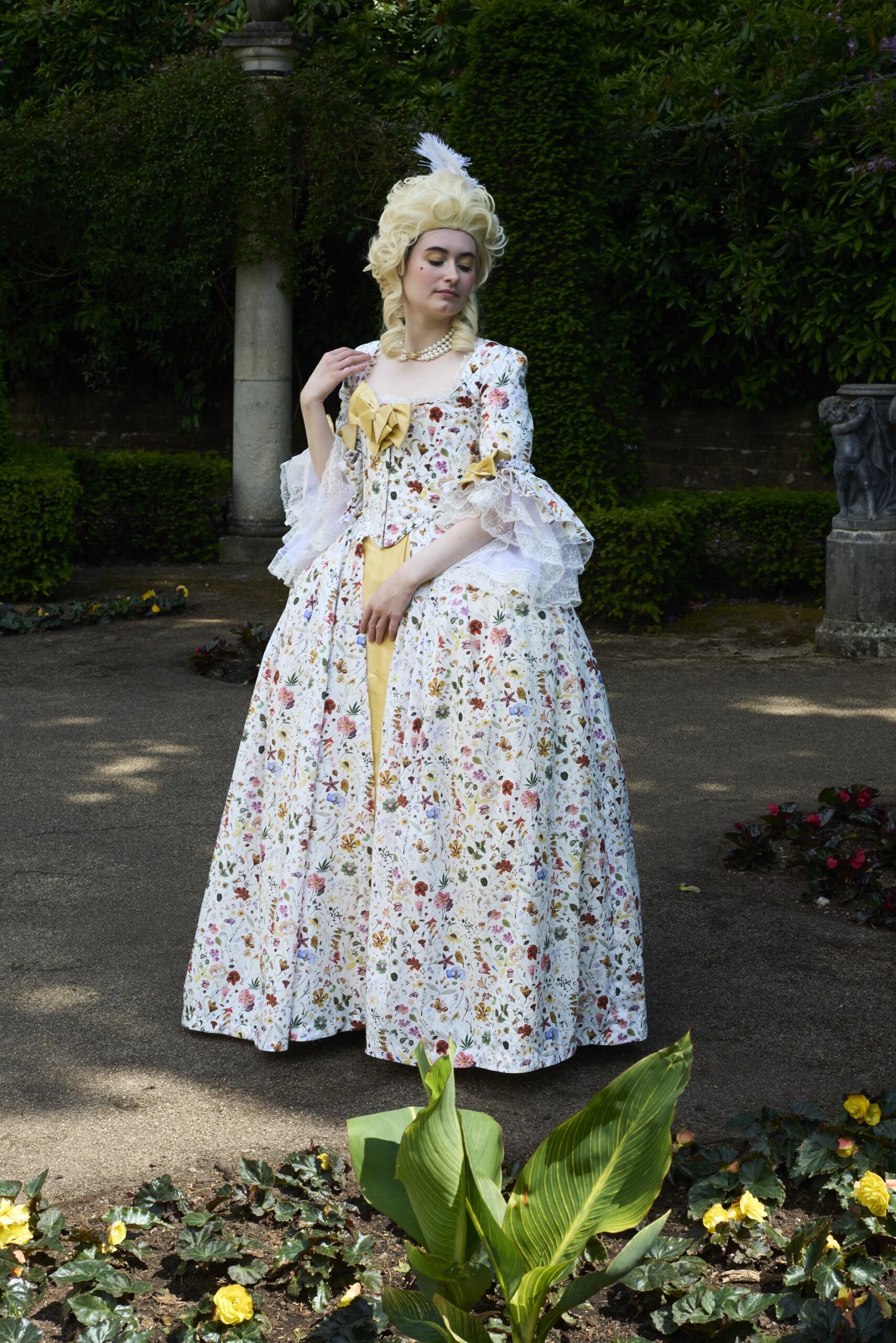 Woman in 18th-century costume with blonde powdered wig and white floral gown standing in formal garden with green foliage backdrop.