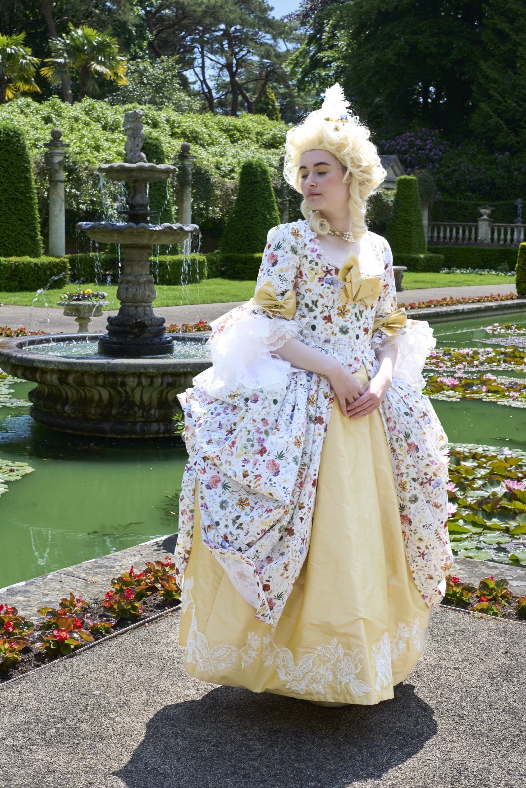 Woman in 18th-century costume with powdered wig sits beside ornate fountain in formal garden with trimmed hedges and lily pond.