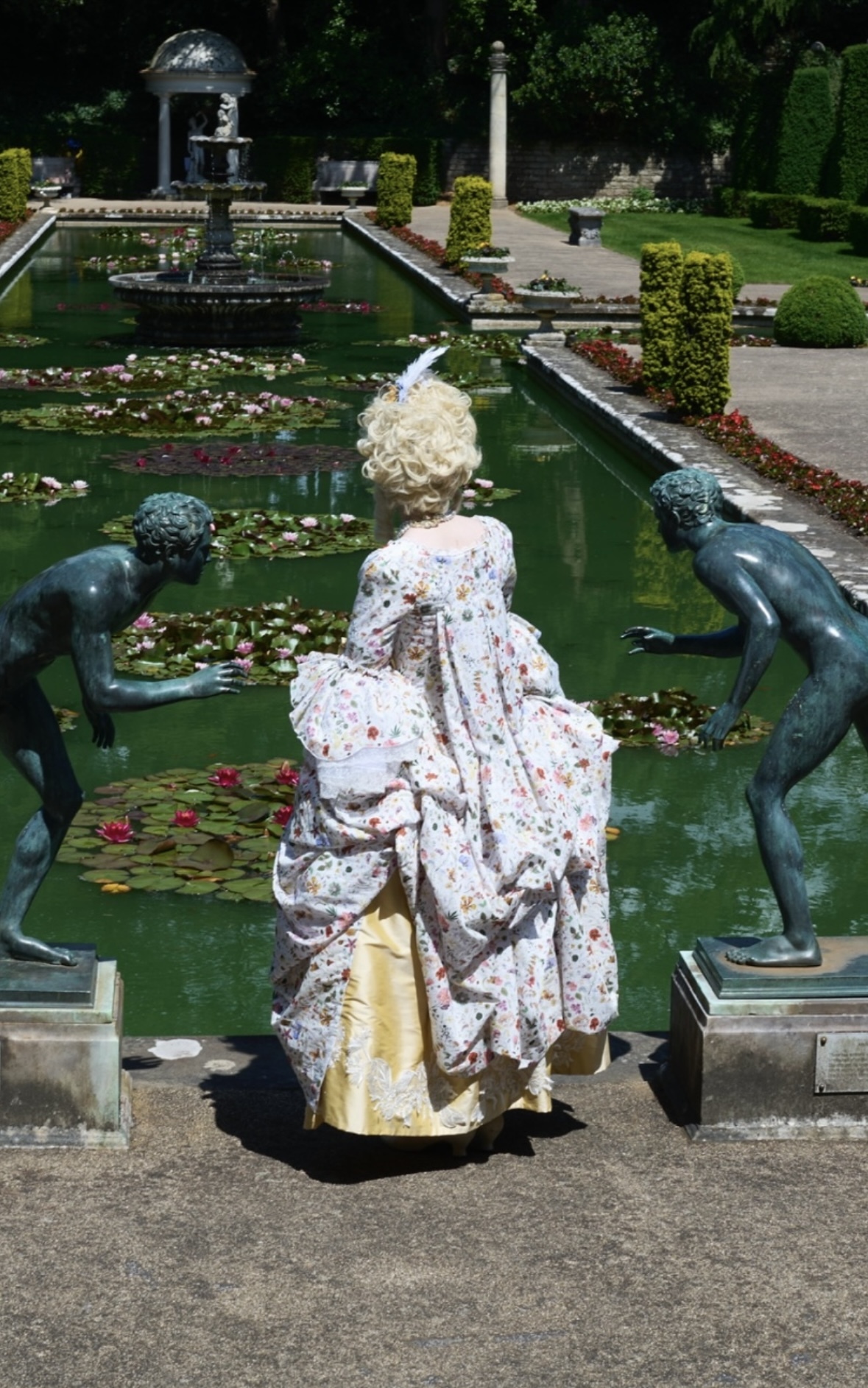 Person in ornate floral dress and blonde wig sits by formal pond with bronze statues and lily pads in landscaped garden.
