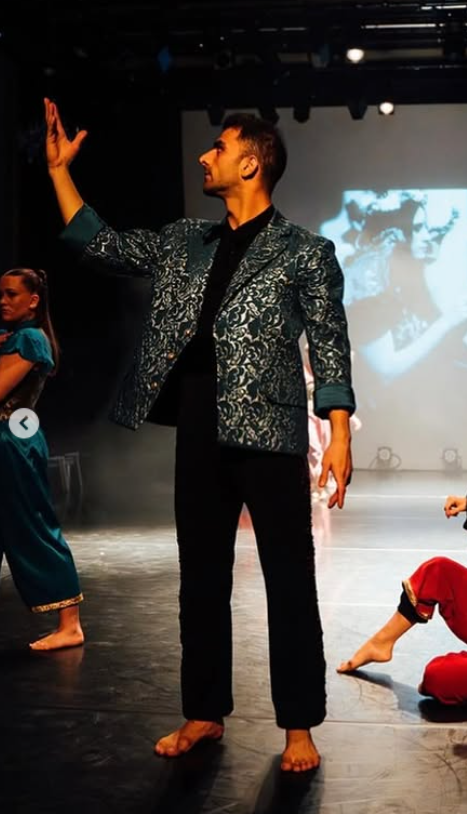 Man in metallic patterned jacket and black trousers gesturing with raised arm on stage, with projection screen and other performers visible.