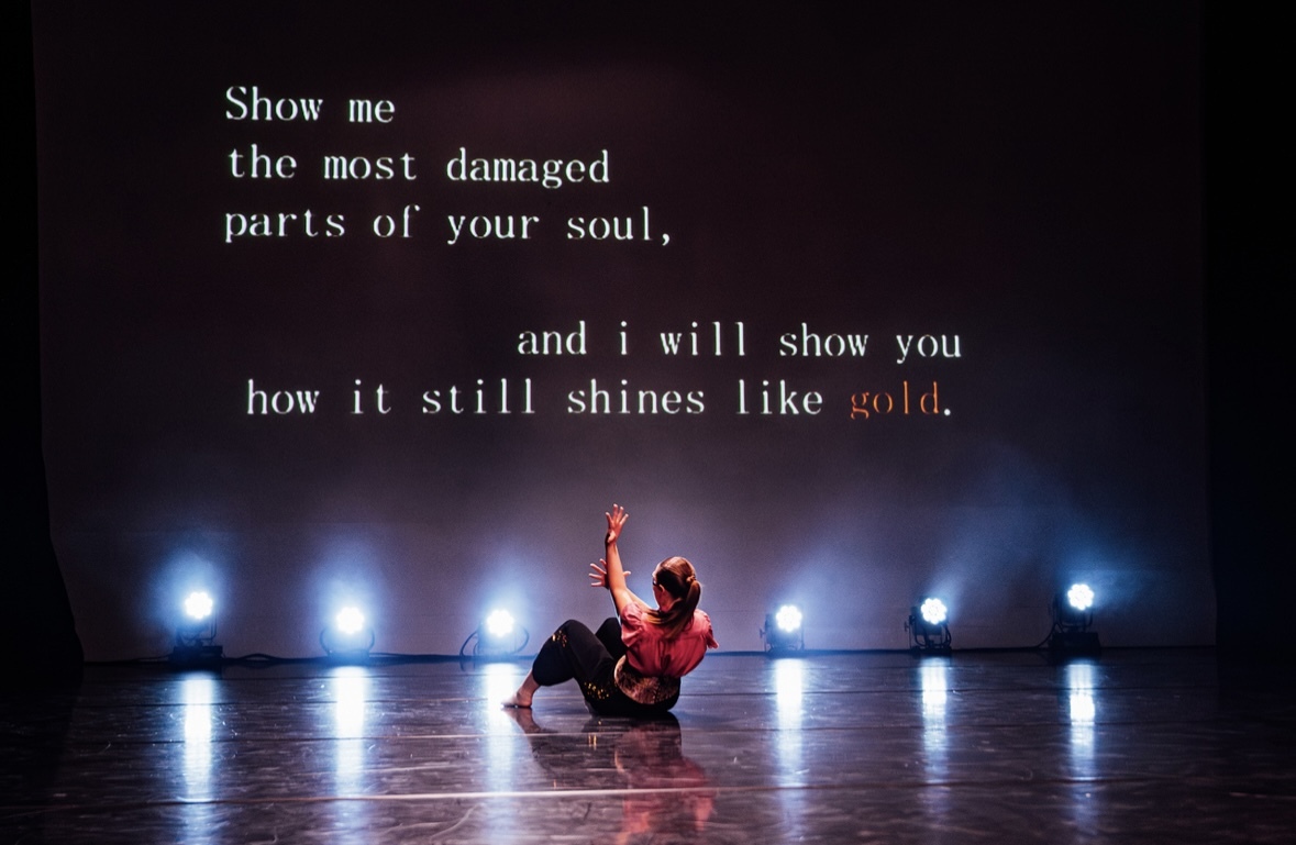 Person kneeling on reflective floor with one arm raised, silhouetted against bright blue stage lights. White text overlay on dark background.